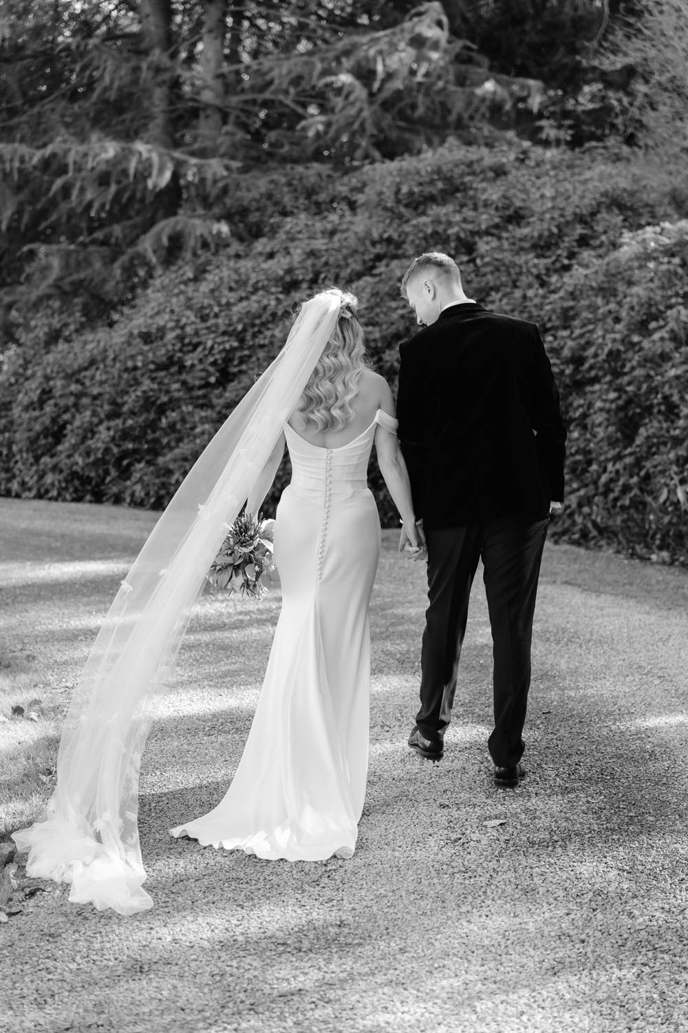 Bride in fitted gown with long lace veil walks with groom through garden path.