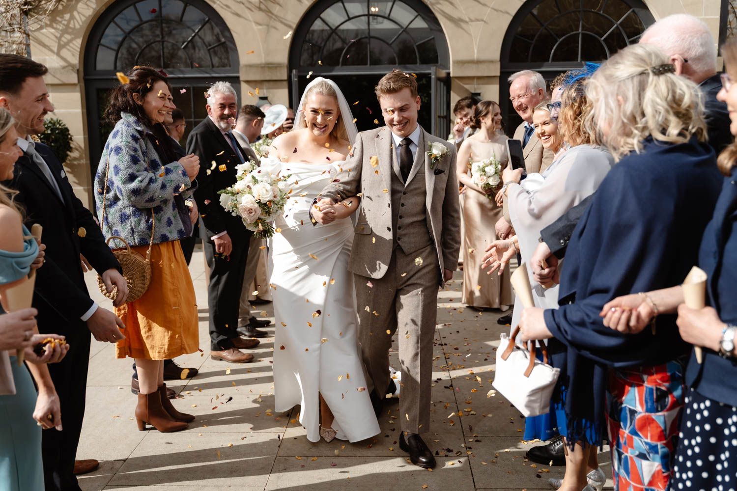 Newlyweds walking through confetti shower outside elegant venue surrounded by cheering guests.