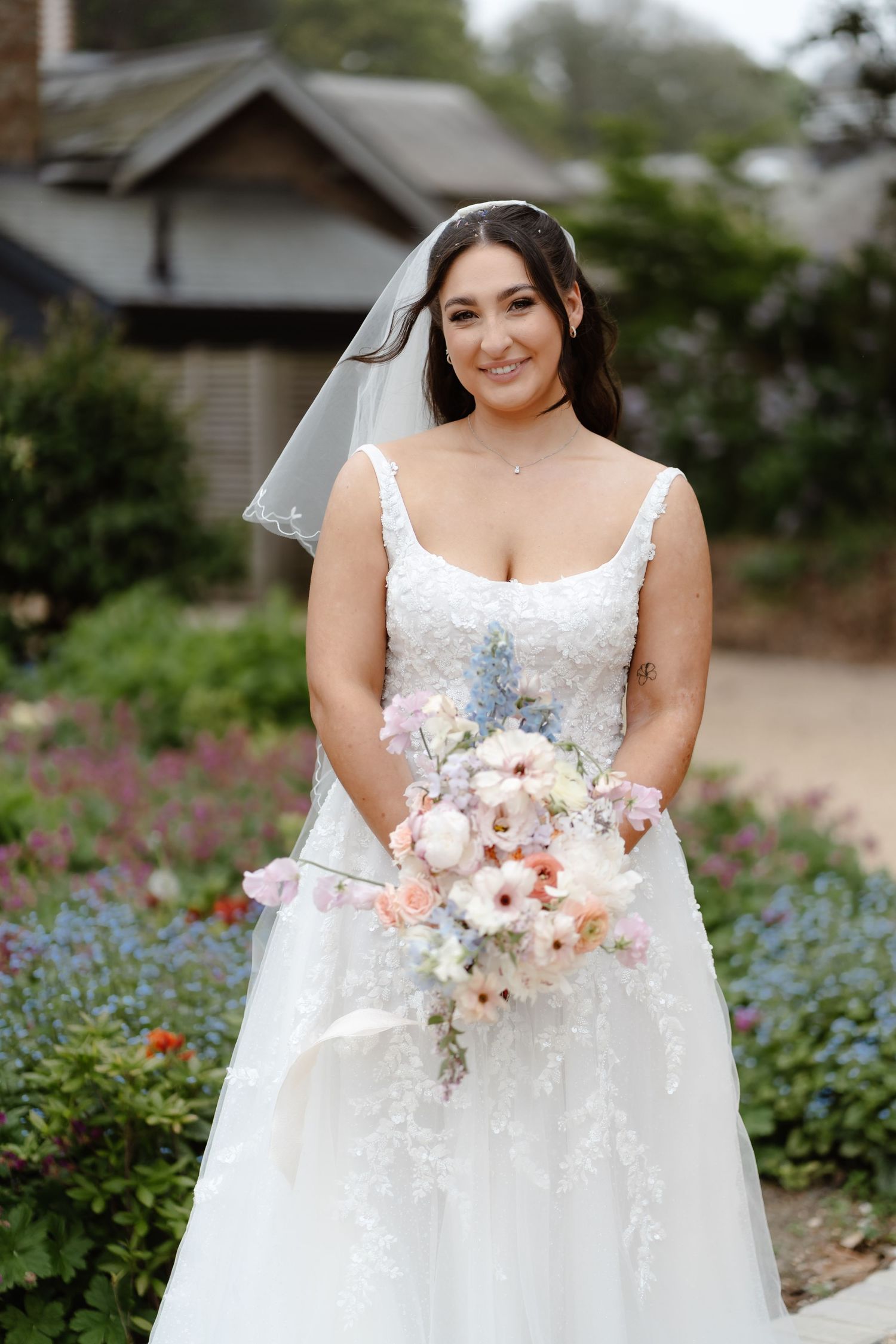 Smiling bride in white lace gown and veil holds pastel bouquet of pink and blue flowers in a garden setting.