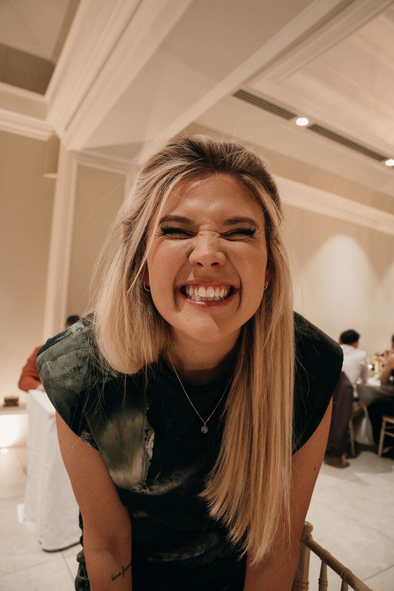 Cheerful blonde woman making a fun expression while leaning forward at an elegant indoor venue.
