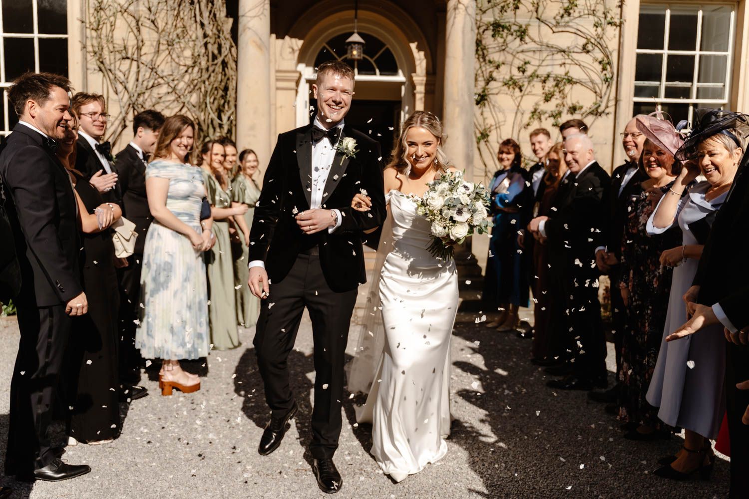 Bride and groom walk through confetti shower with smiling wedding guests lining the path outside elegant venue.