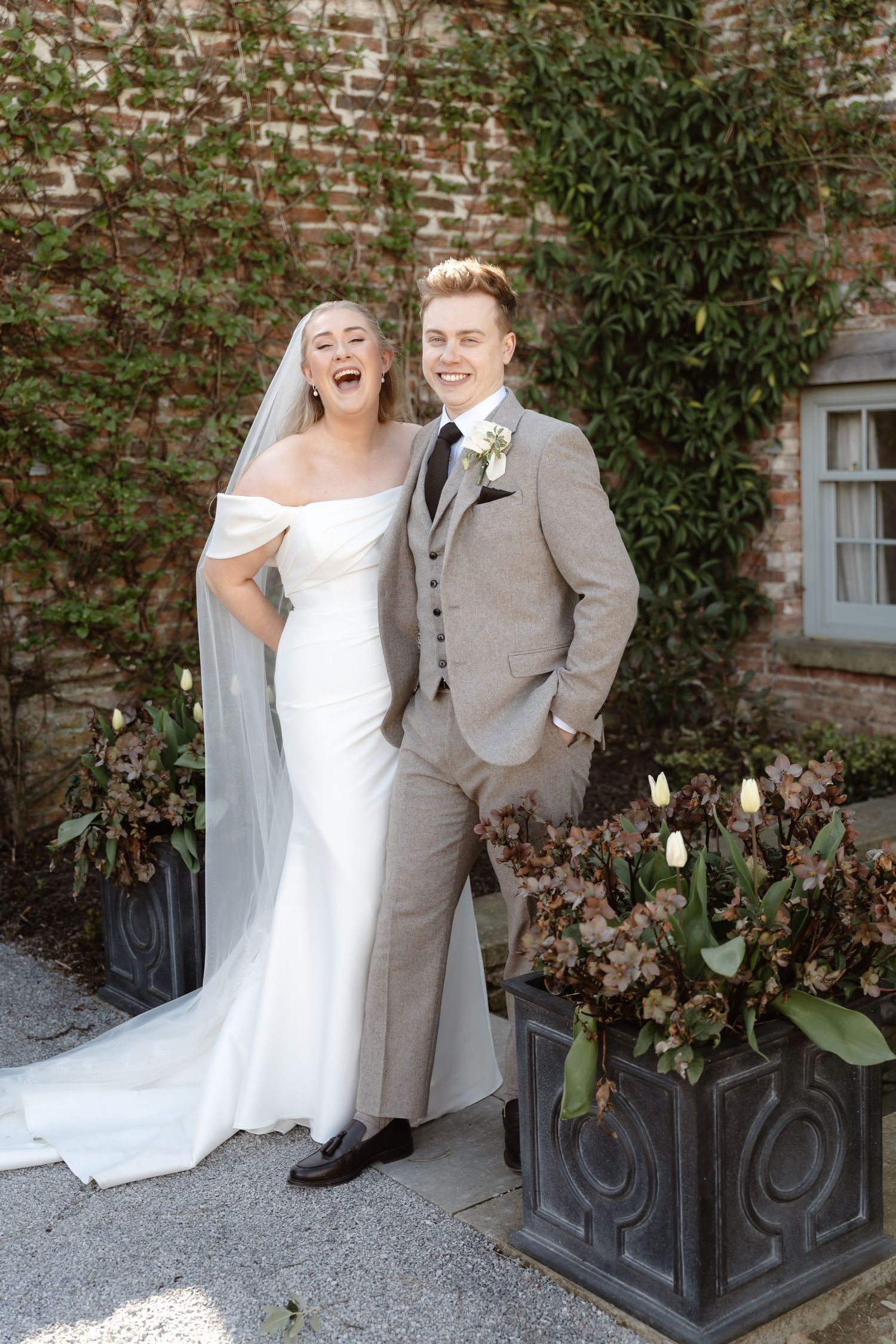 Laughing bride in white gown and veil poses with smiling groom in grey tweed suit outside brick building.