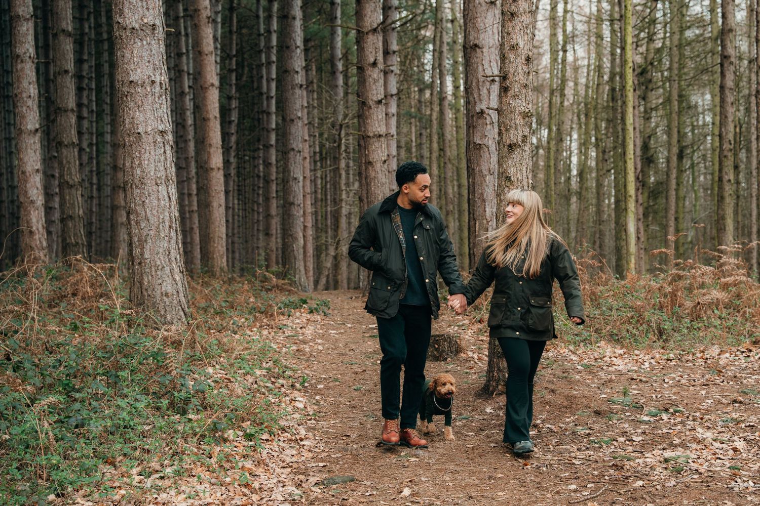 Smiling couple holding hands walking along a woodland path with their small dog.
