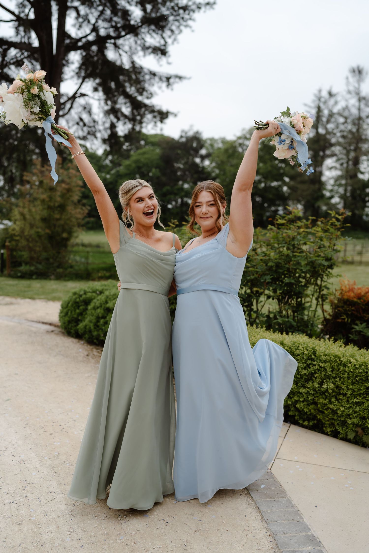 Two bridesmaids in sage green and blue gowns joyfully raise bouquets outdoors at a wedding celebration.
