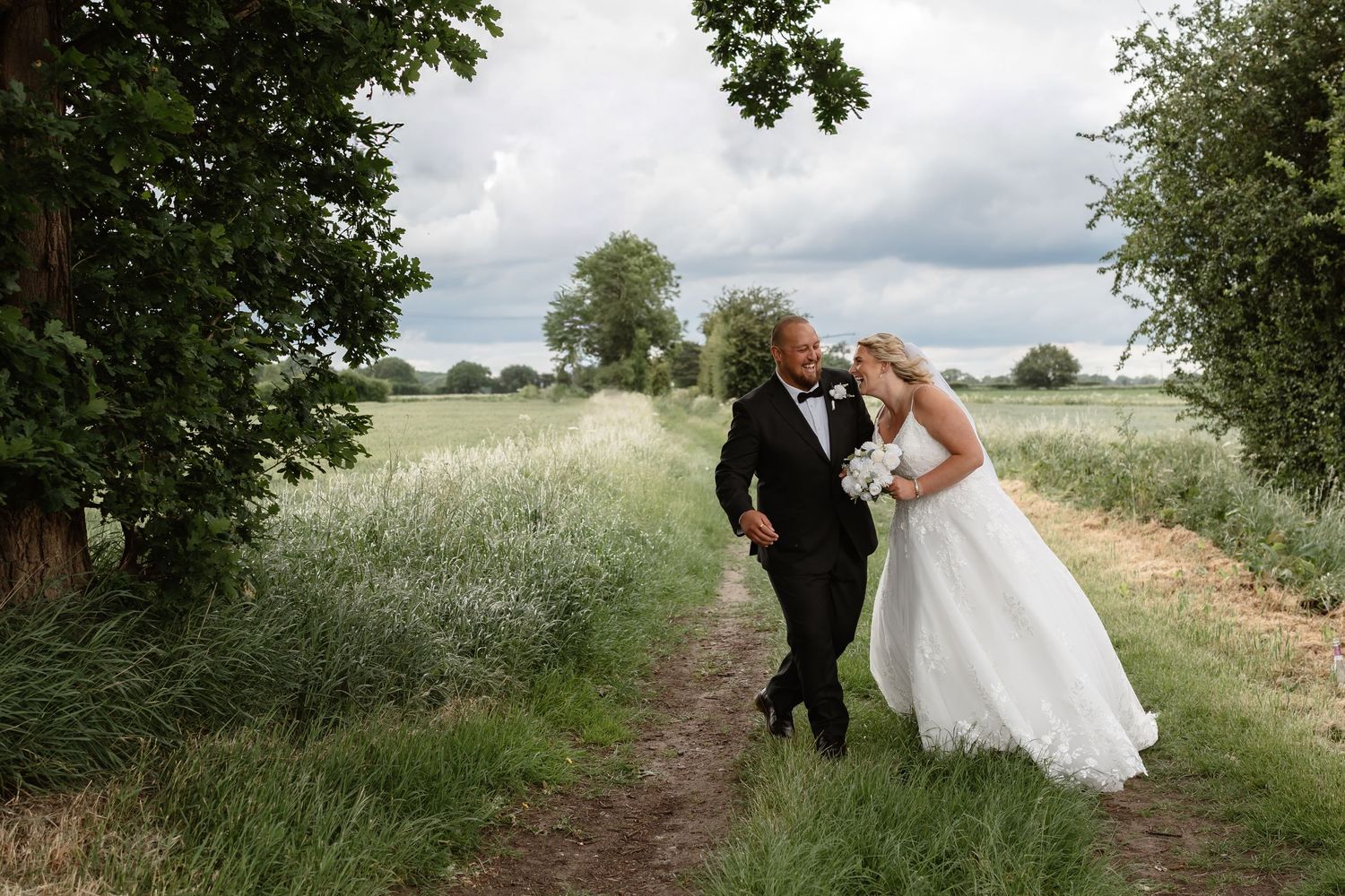 Bride and groom walking hand in hand along a scenic countryside path surrounded by lush green fields on their wedding day.