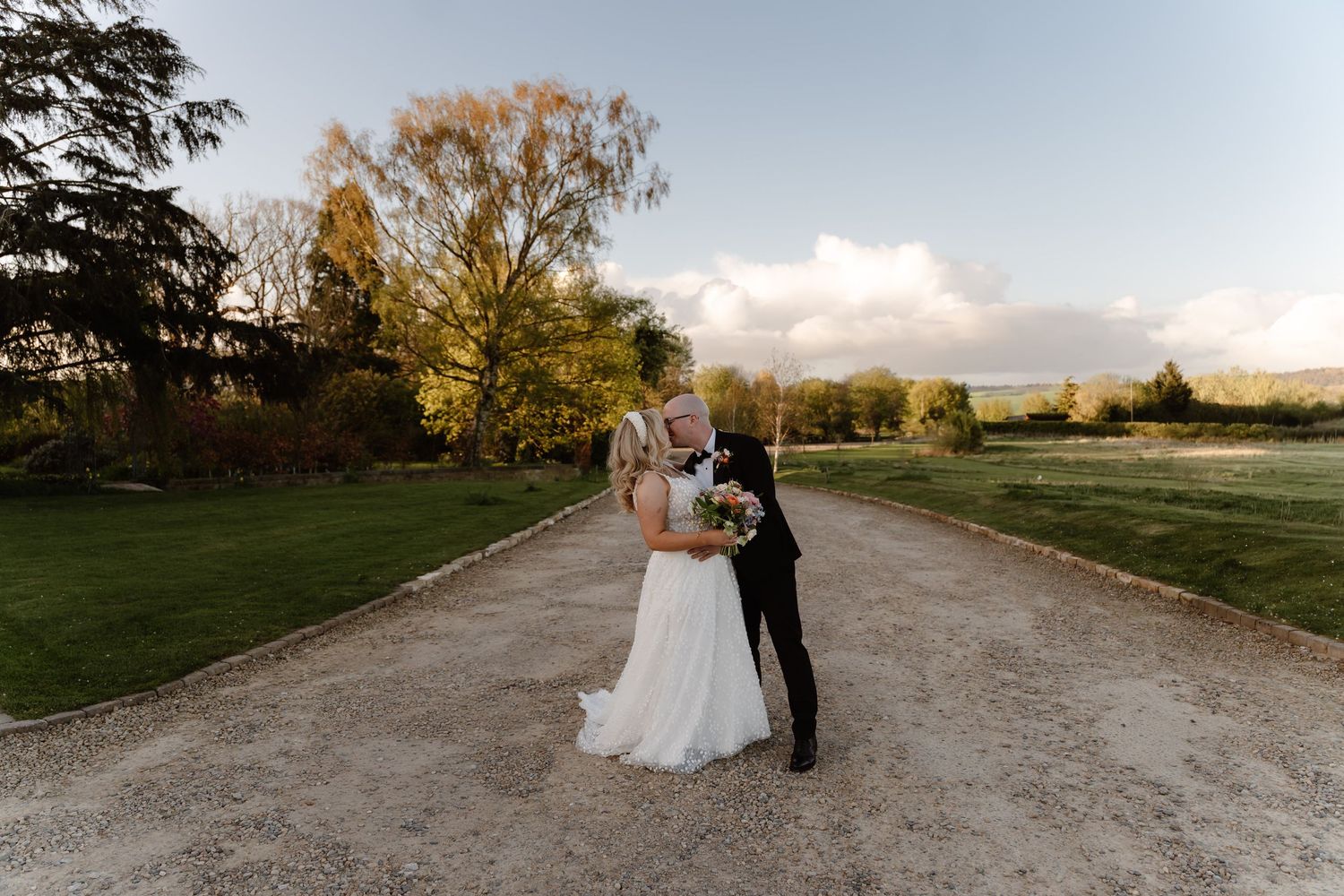 Bride and groom sharing a romantic kiss on a gravel path surrounded by lush greenery at golden hour.
