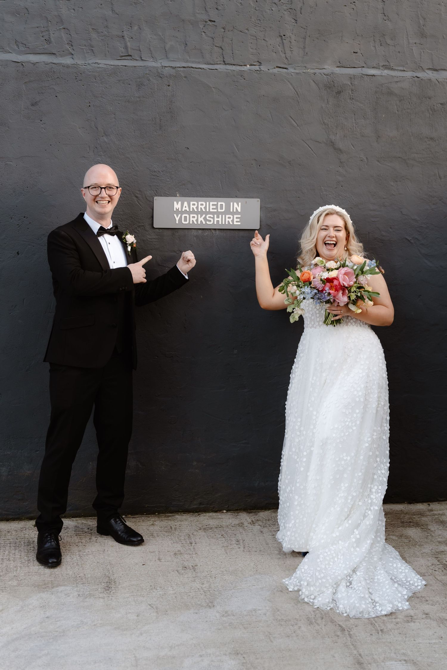 Newlyweds pointing at a Married in Yorkshire sign, bride in white gown holding bouquet, groom in black tuxedo.