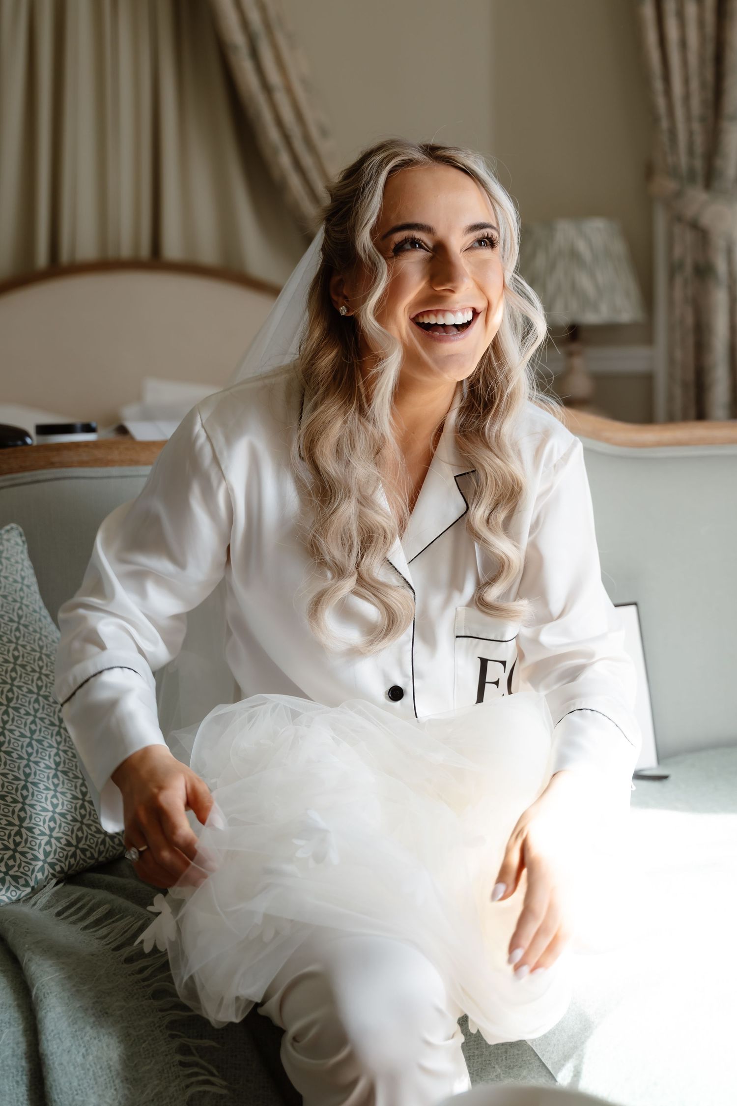 Radiant blonde bride in white monogrammed satin pajamas laughing while holding wedding veil.