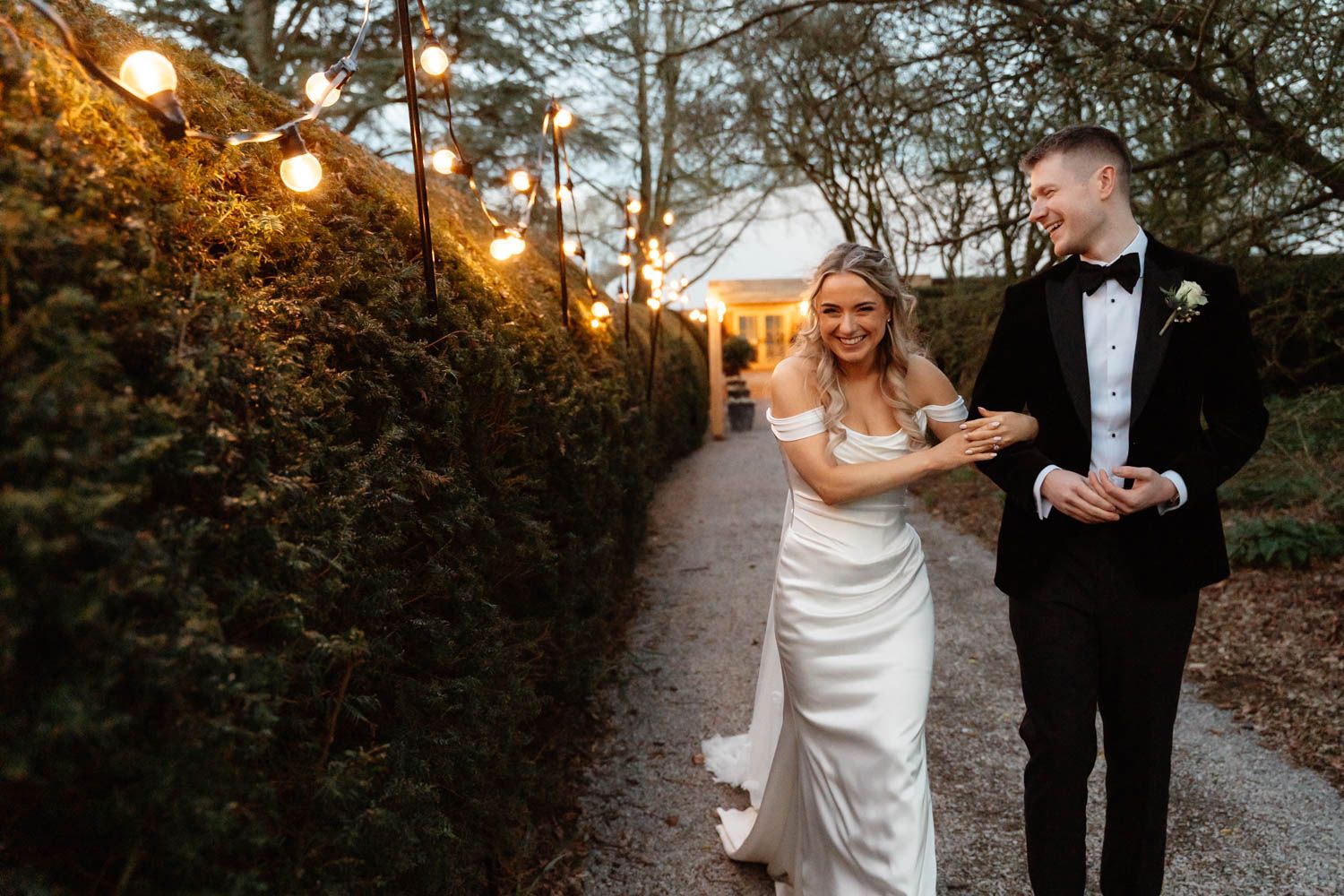Bride and groom laughing together on a garden path lined with hedges and string lights at dusk.
