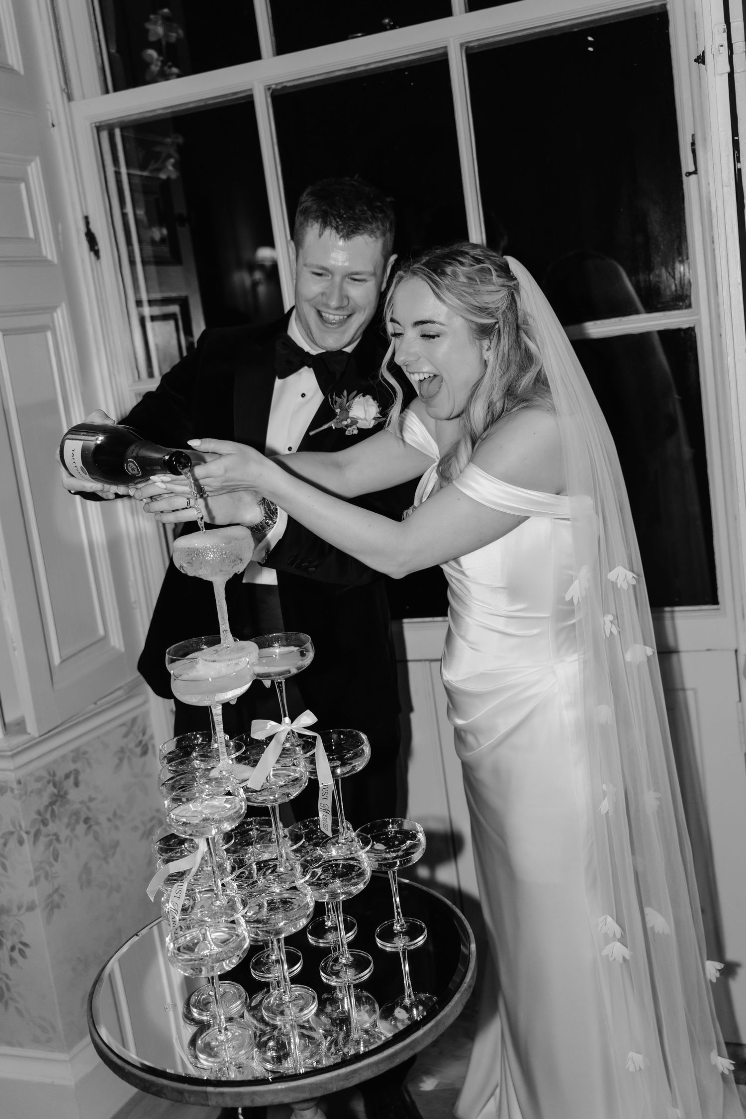 Bride and groom laughing while pouring champagne tower at wedding reception in black and white photo.