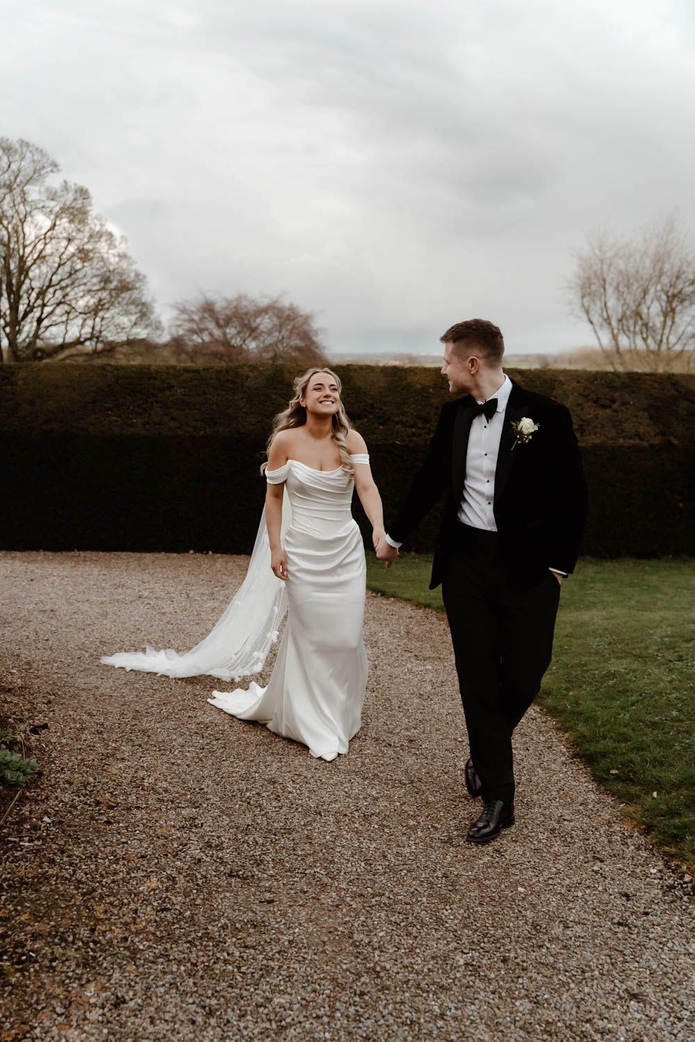 Bride and groom walking hand in hand along gravel path through manicured hedgerow garden.