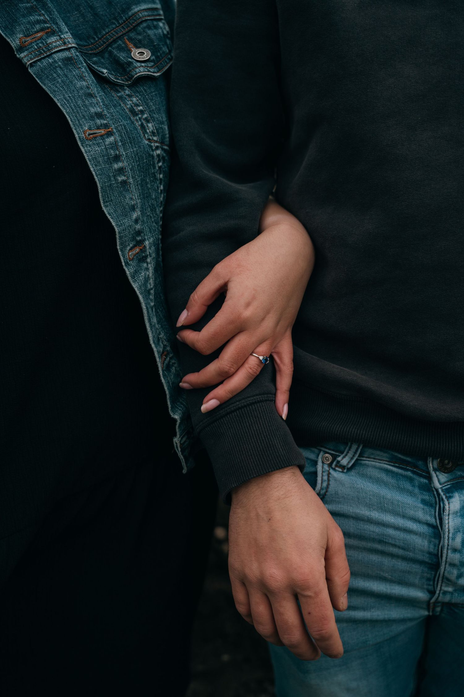 Close-up of couple holding hands, woman wearing engagement ring, man in black sweater and jeans.