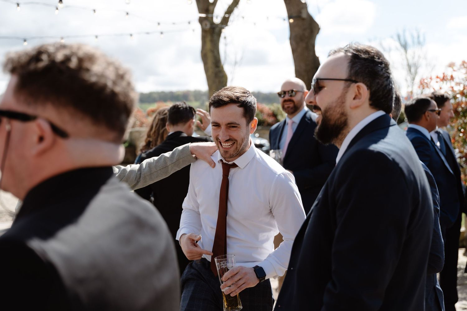Men in suits socializing outdoors at a wedding reception, smiling and enjoying drinks in a garden setting.