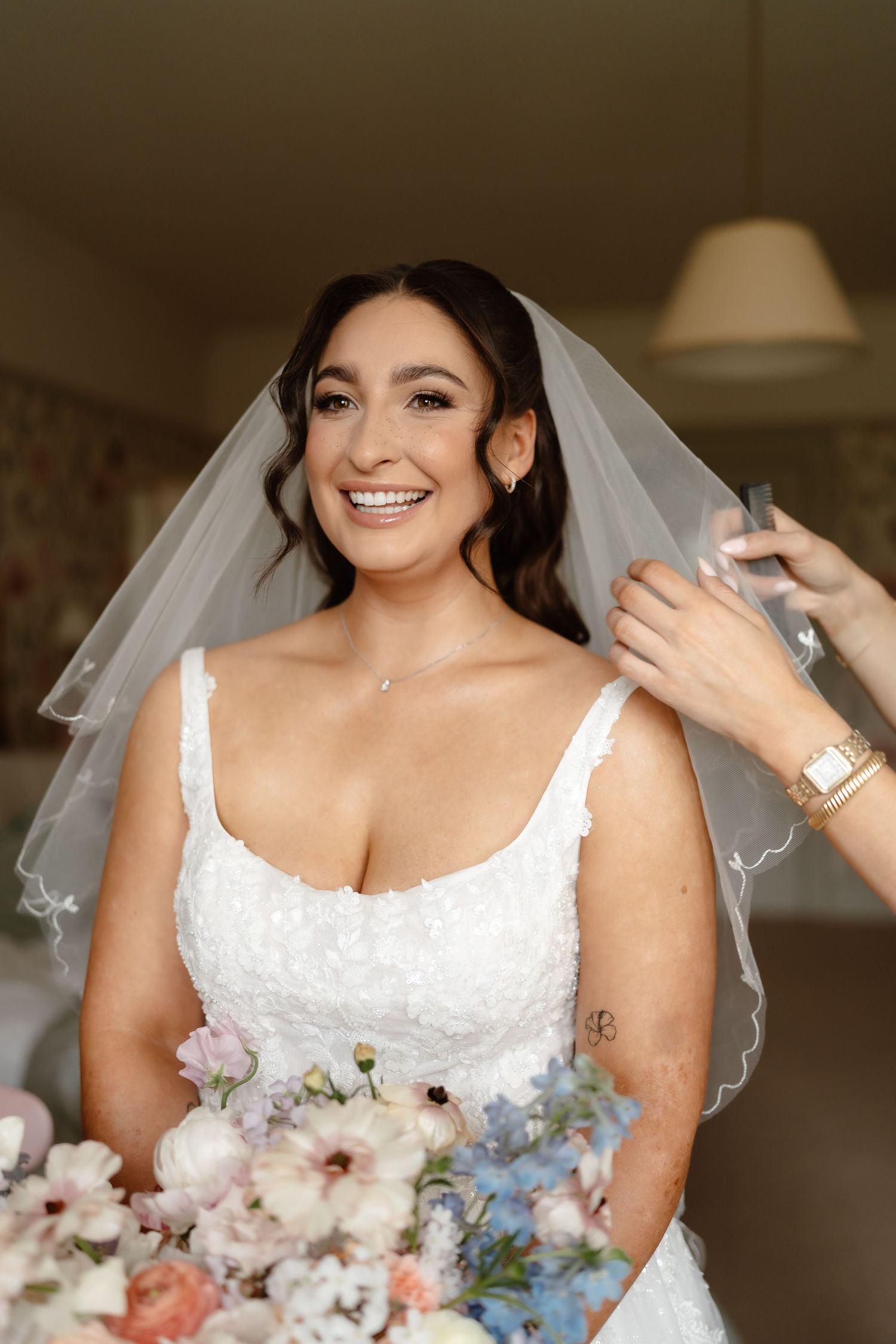 Smiling bride in white lace gown and veil holds pastel bouquet while getting ready indoors.