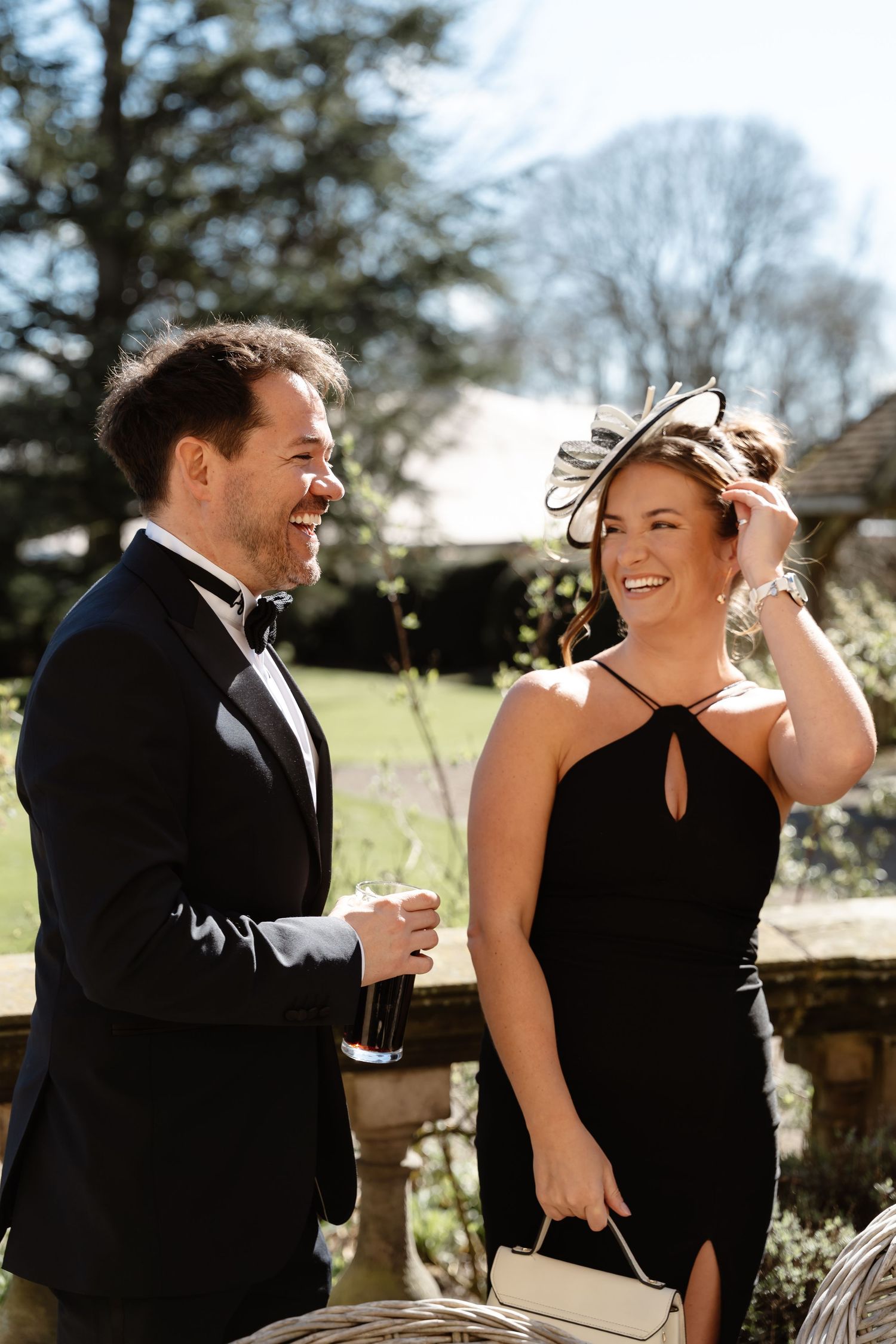 Elegantly dressed wedding guests in black tie attire laughing outdoors in bright spring sunshine.