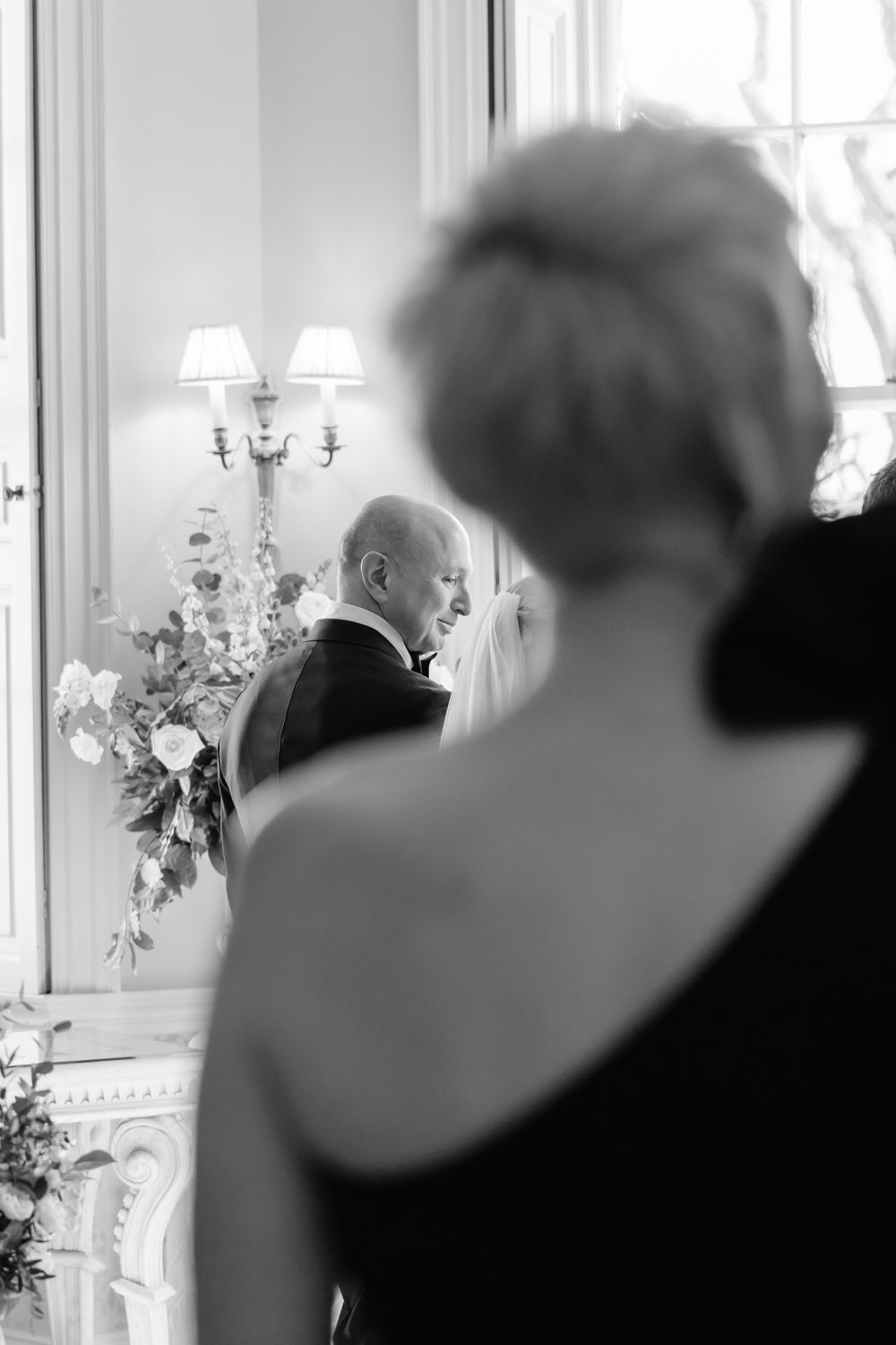 Black and white photo of wedding guests watching the ceremony with floral arrangements and wall sconces in background.
