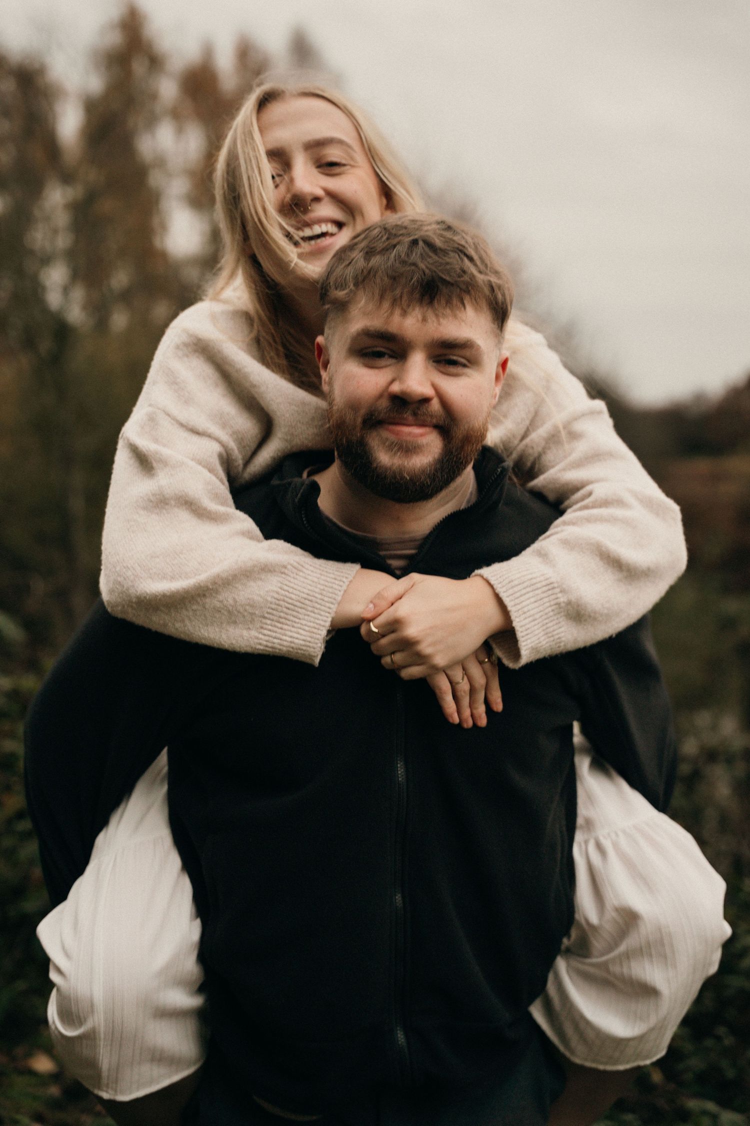 Smiling couple posing outdoors, woman giving man a playful embrace from behind in an autumn setting.