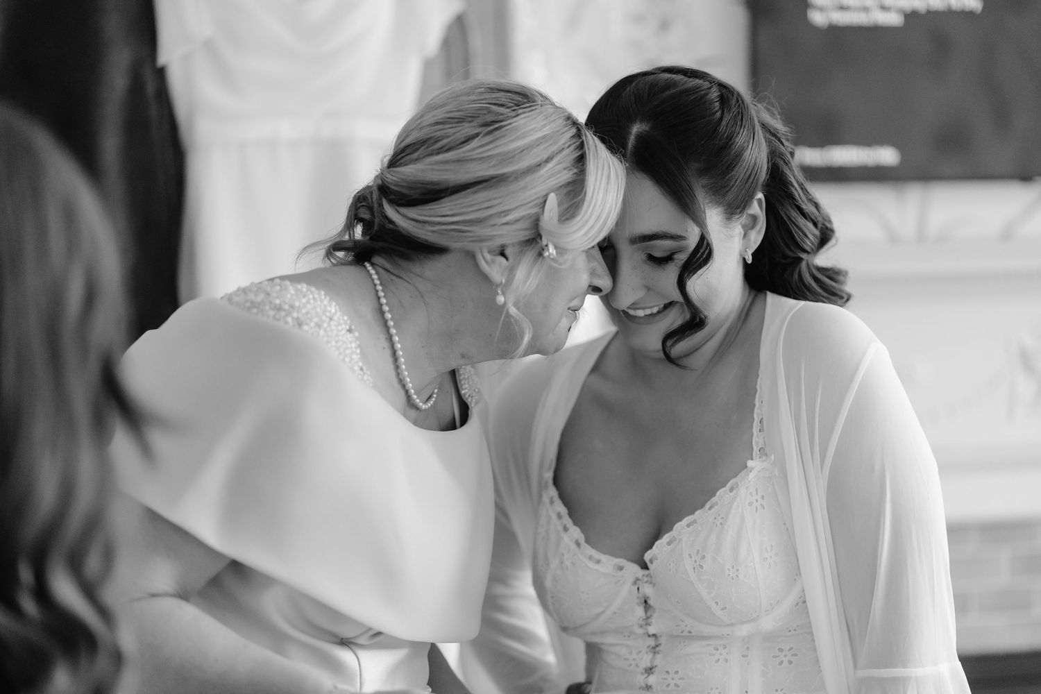 Black and white photo of a bride smiling as an older woman whispers to her during wedding preparations.