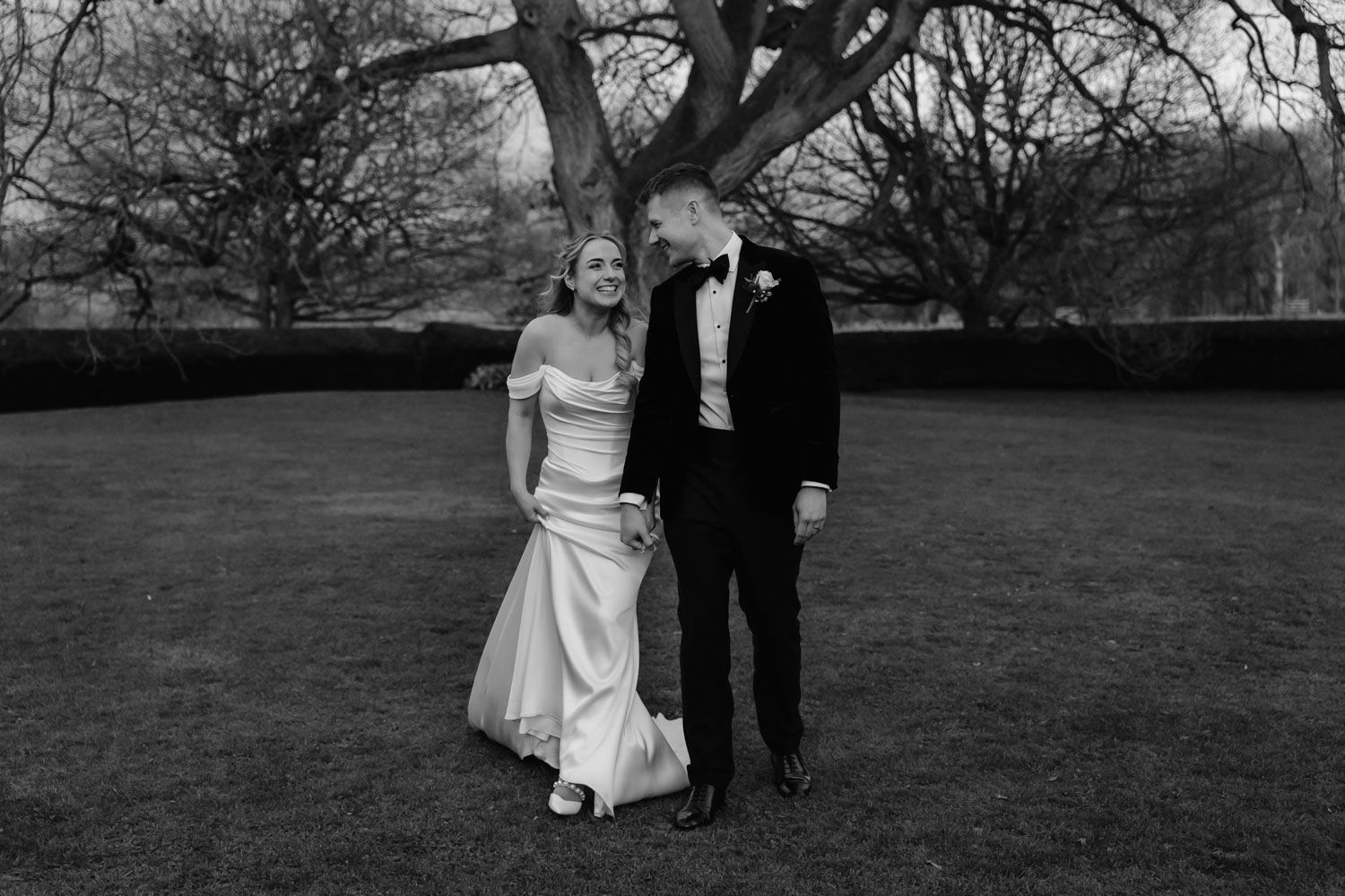 Bride and groom share a joyful moment on a lawn in black and white, bride in off-shoulder gown, groom in black tuxedo.