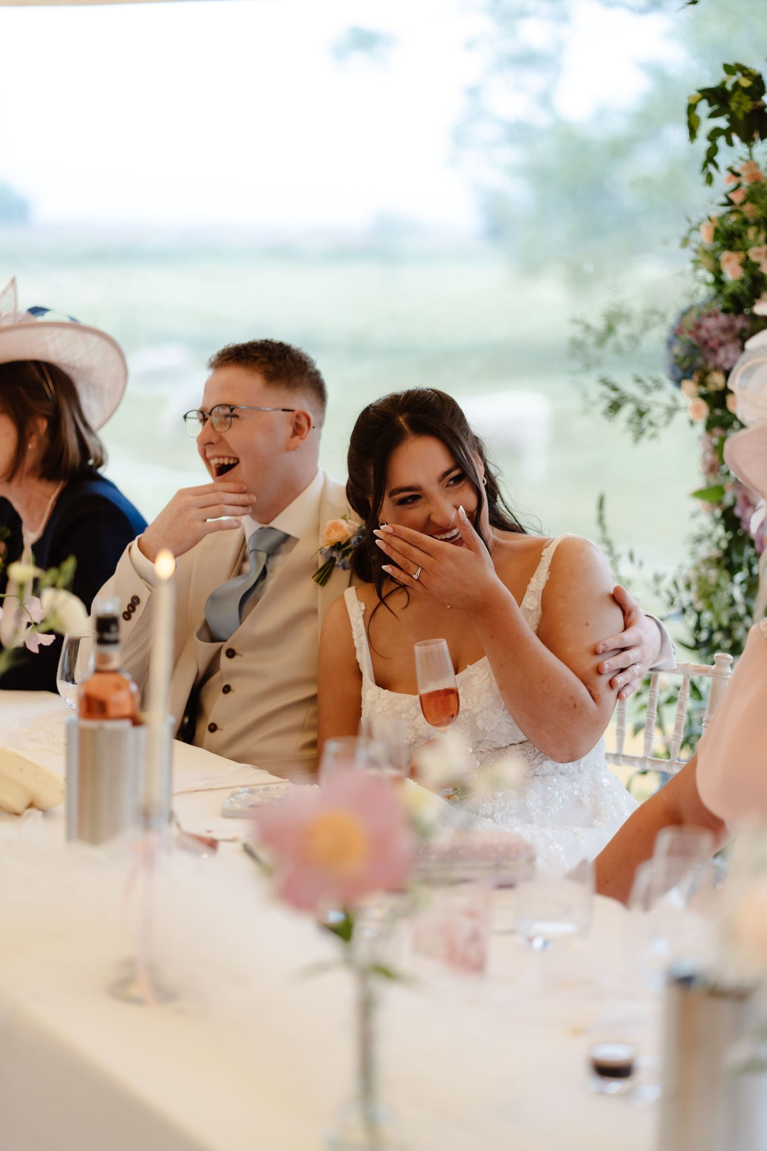 Bride and groom laughing joyfully during wedding reception speeches at elegantly decorated table with floral arrangements.