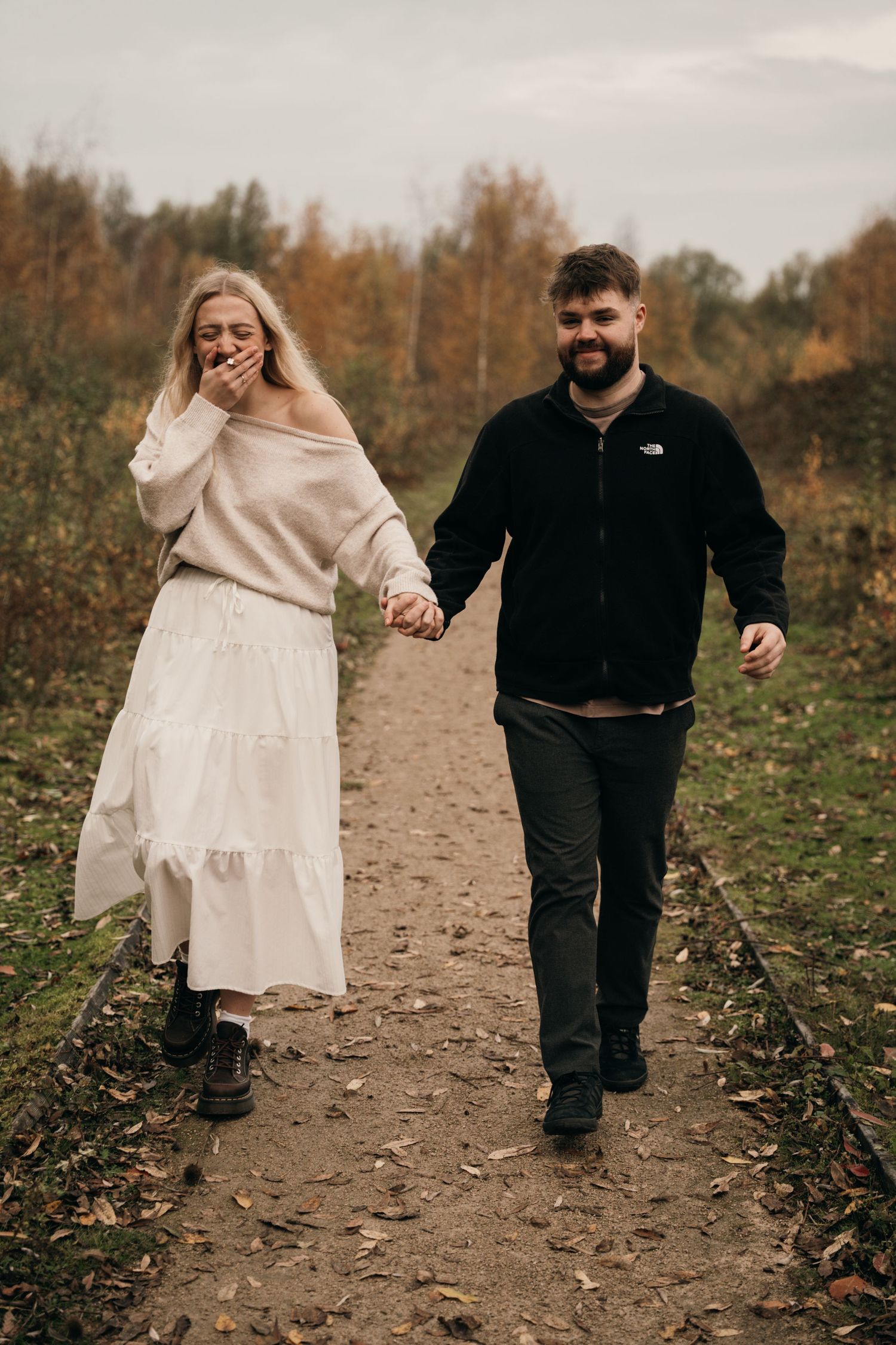 Happy couple holding hands walking on autumn path, woman in white dress and man in black fleece jacket.