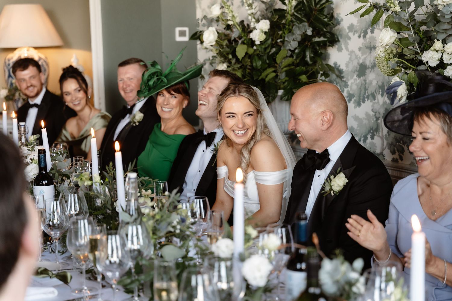 Bride laughs joyfully at wedding reception table surrounded by family, with candles and white floral centerpieces.