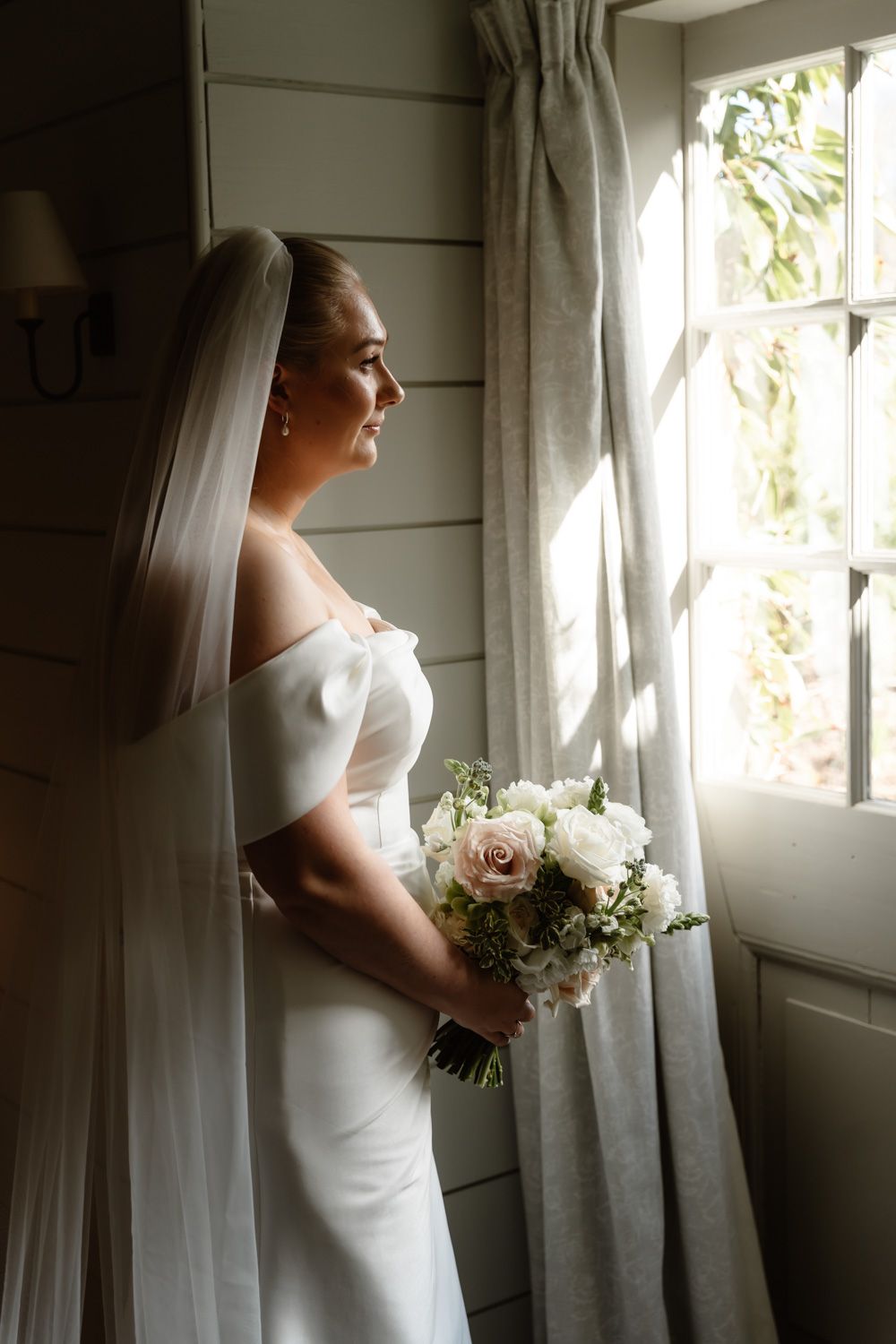 Elegant bride in off-shoulder gown holding white bouquet gazing out sunlit window with veil.