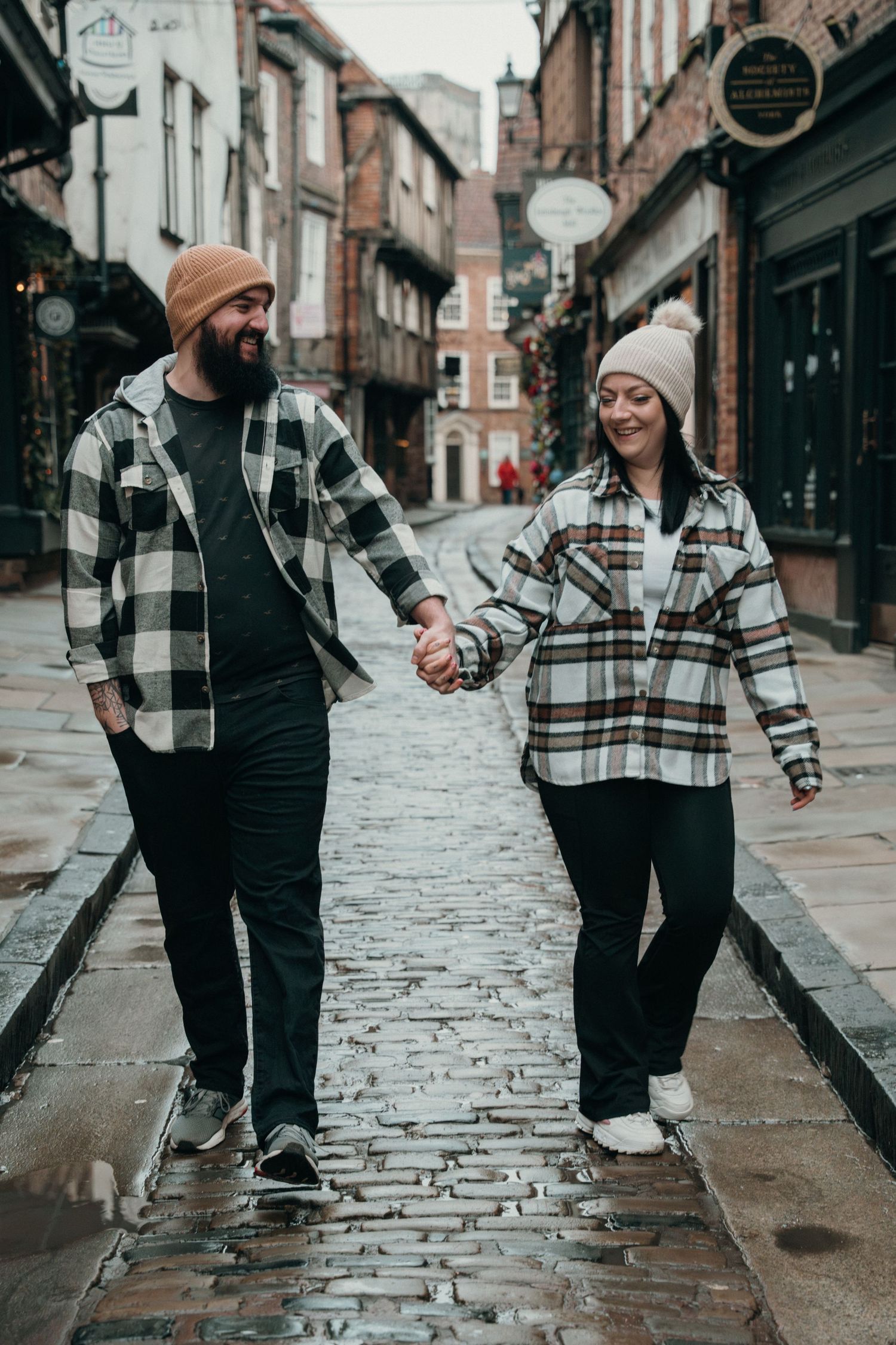 A couple in coordinating plaid shirts hold hands while walking on a wet cobblestone street in York.