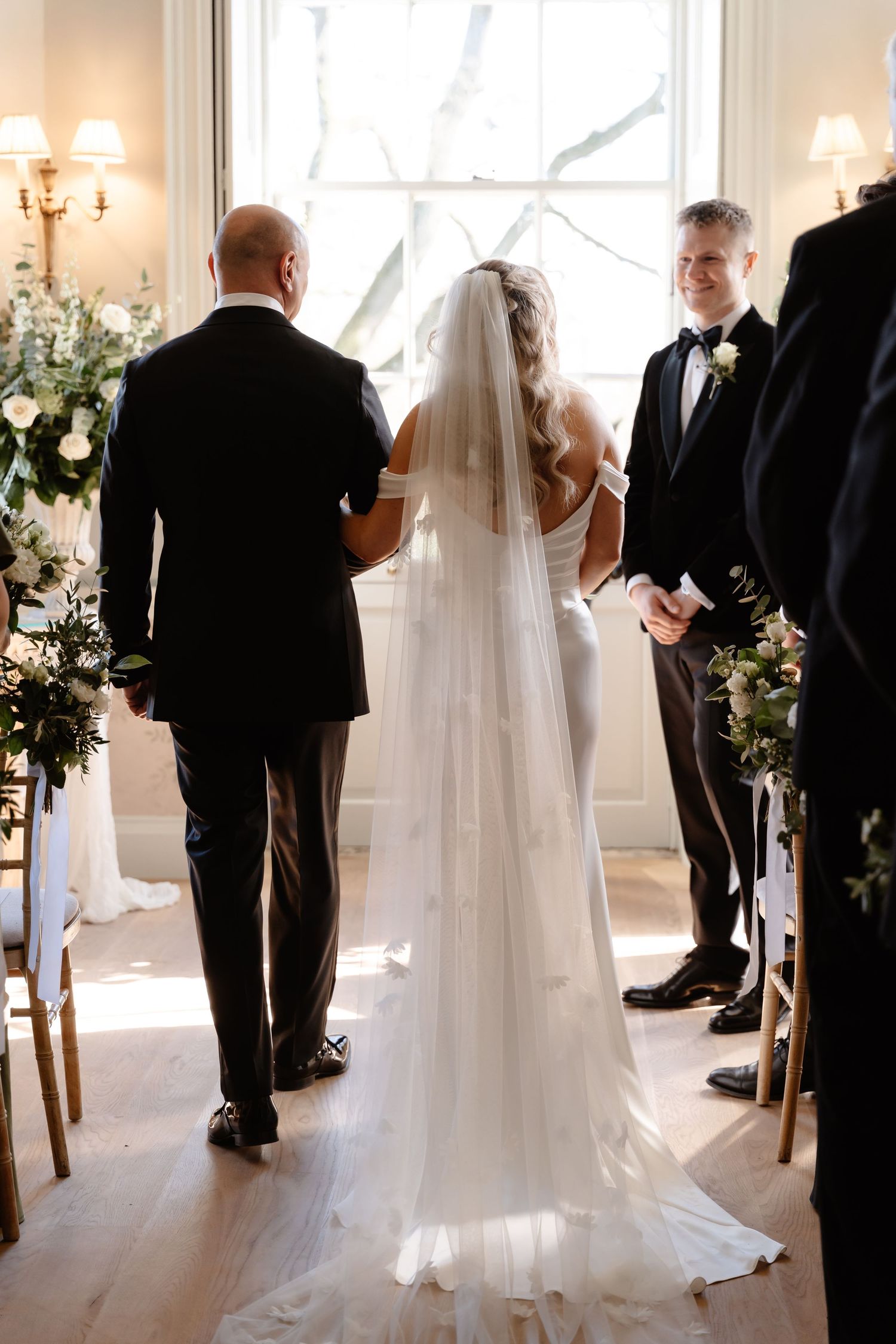 Bride being walked down the aisle by father toward smiling groom at indoor ceremony.