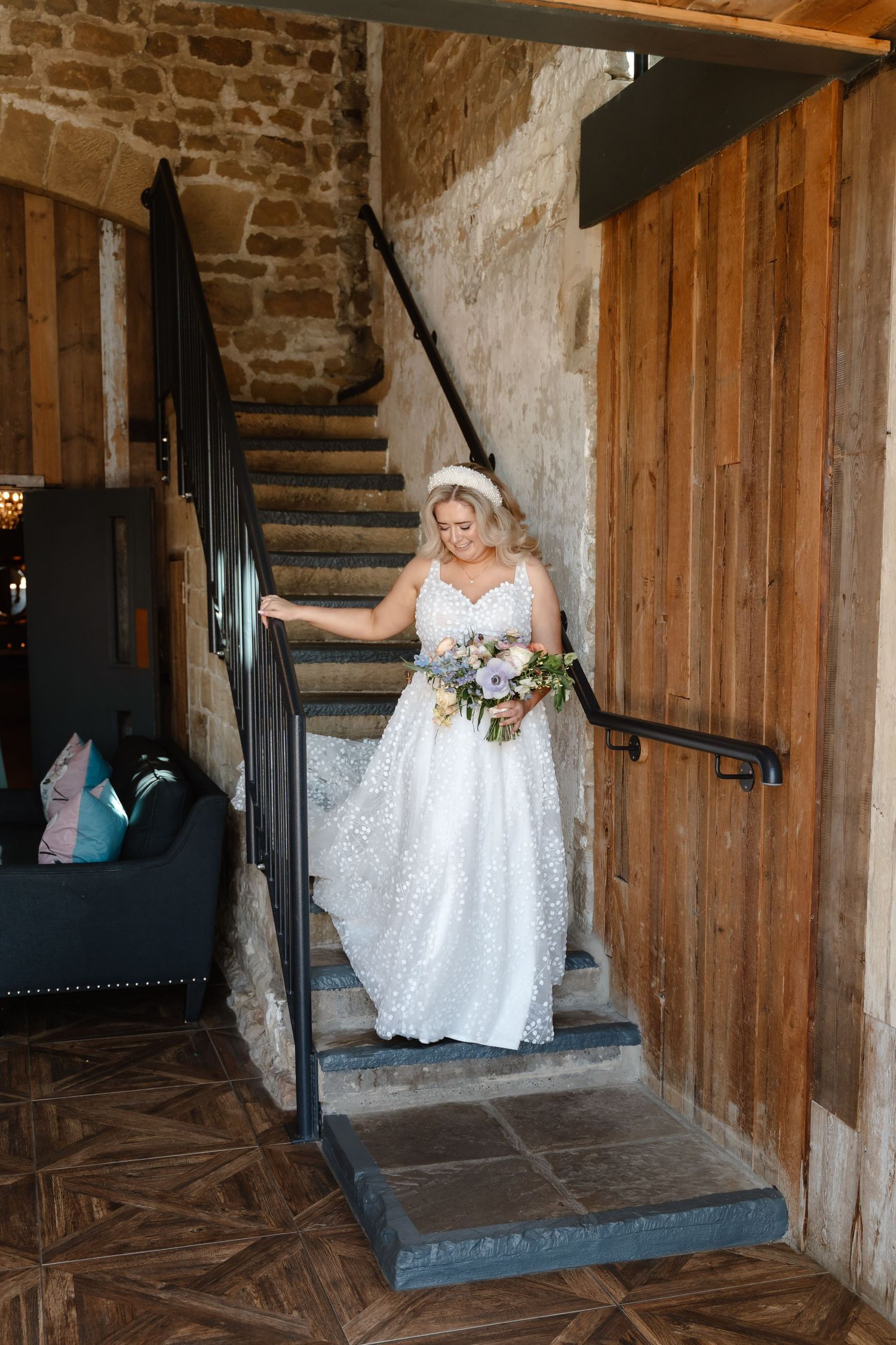 Bride in lace wedding gown descending stone staircase holding floral bouquet in rustic venue.