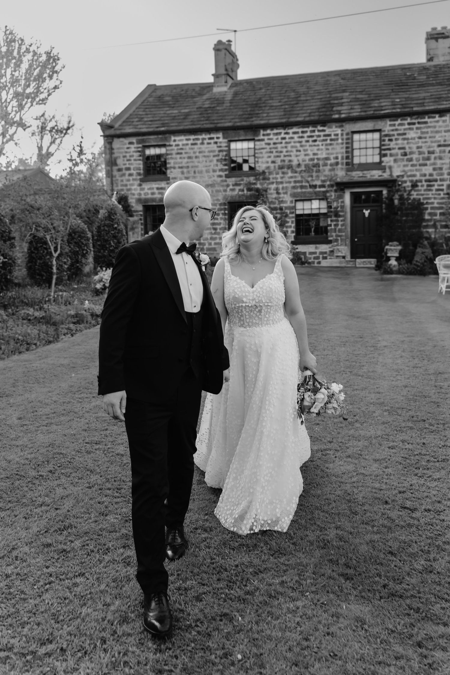 Black and white photo of a bride and groom walking hand in hand outside a stone building on their wedding day.