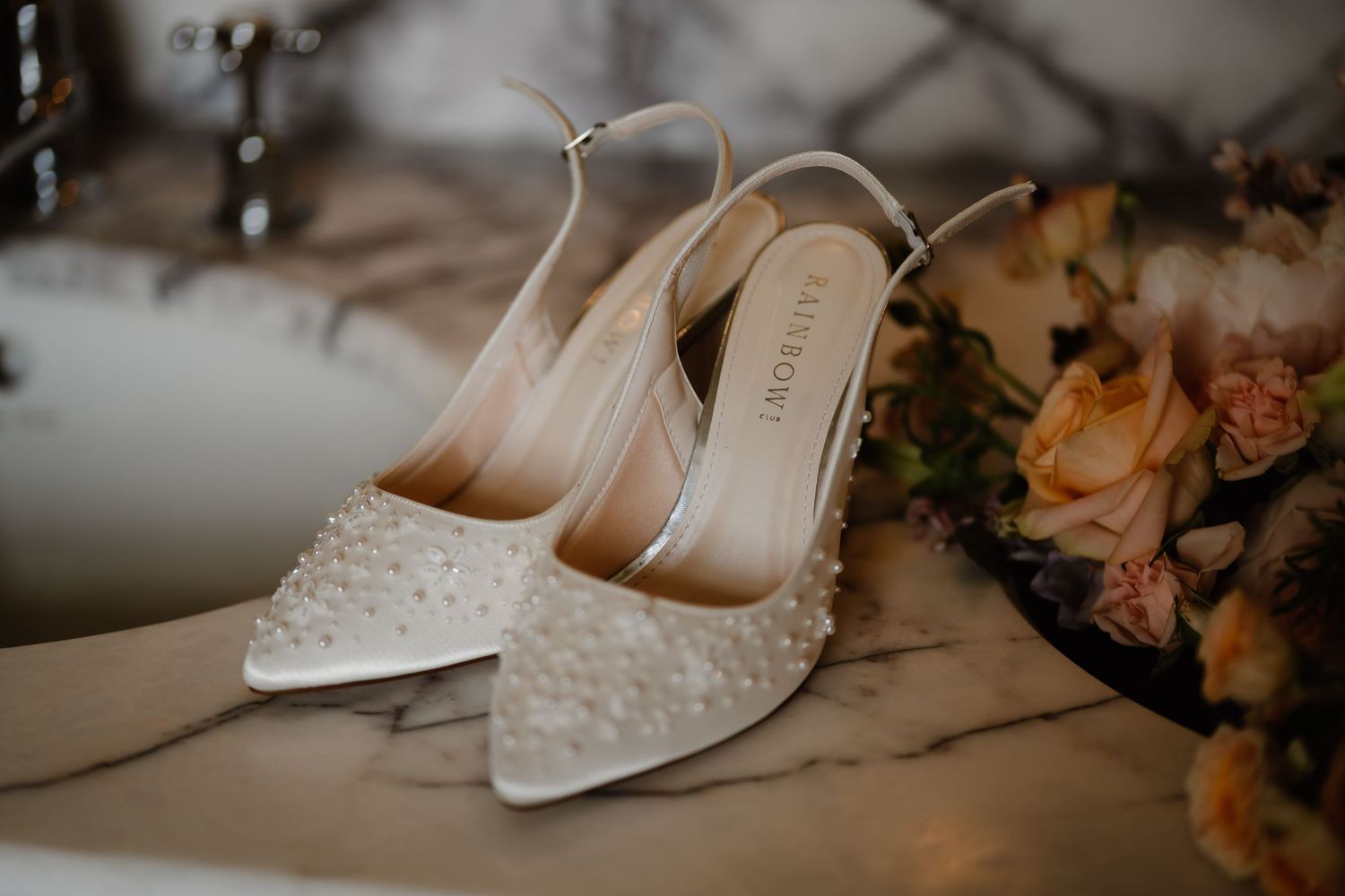 Elegant pearl-embellished ivory slingback wedding heels displayed on marble surface beside a floral bouquet.