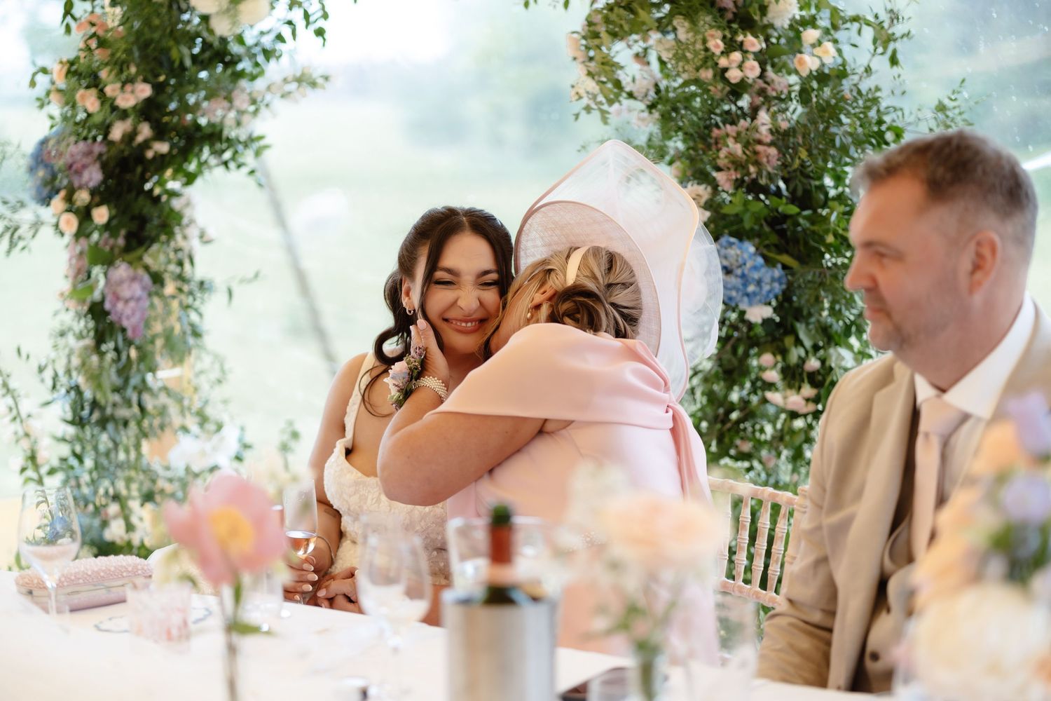 Bride in white gown hugging a guest in a pink hat at an elegant floral wedding reception.