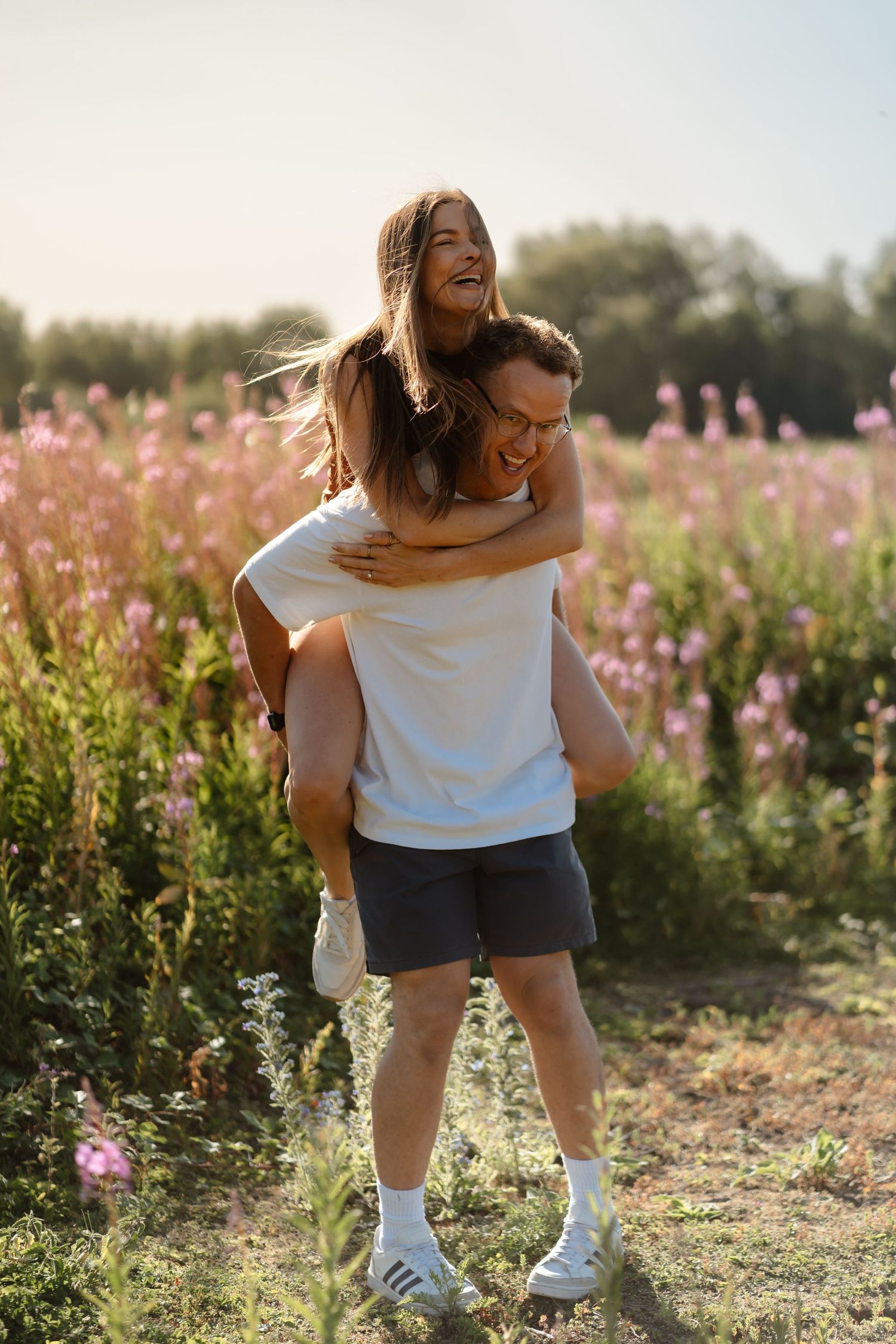 Happy couple enjoying piggyback ride in blooming pink flower field on sunny summer day.