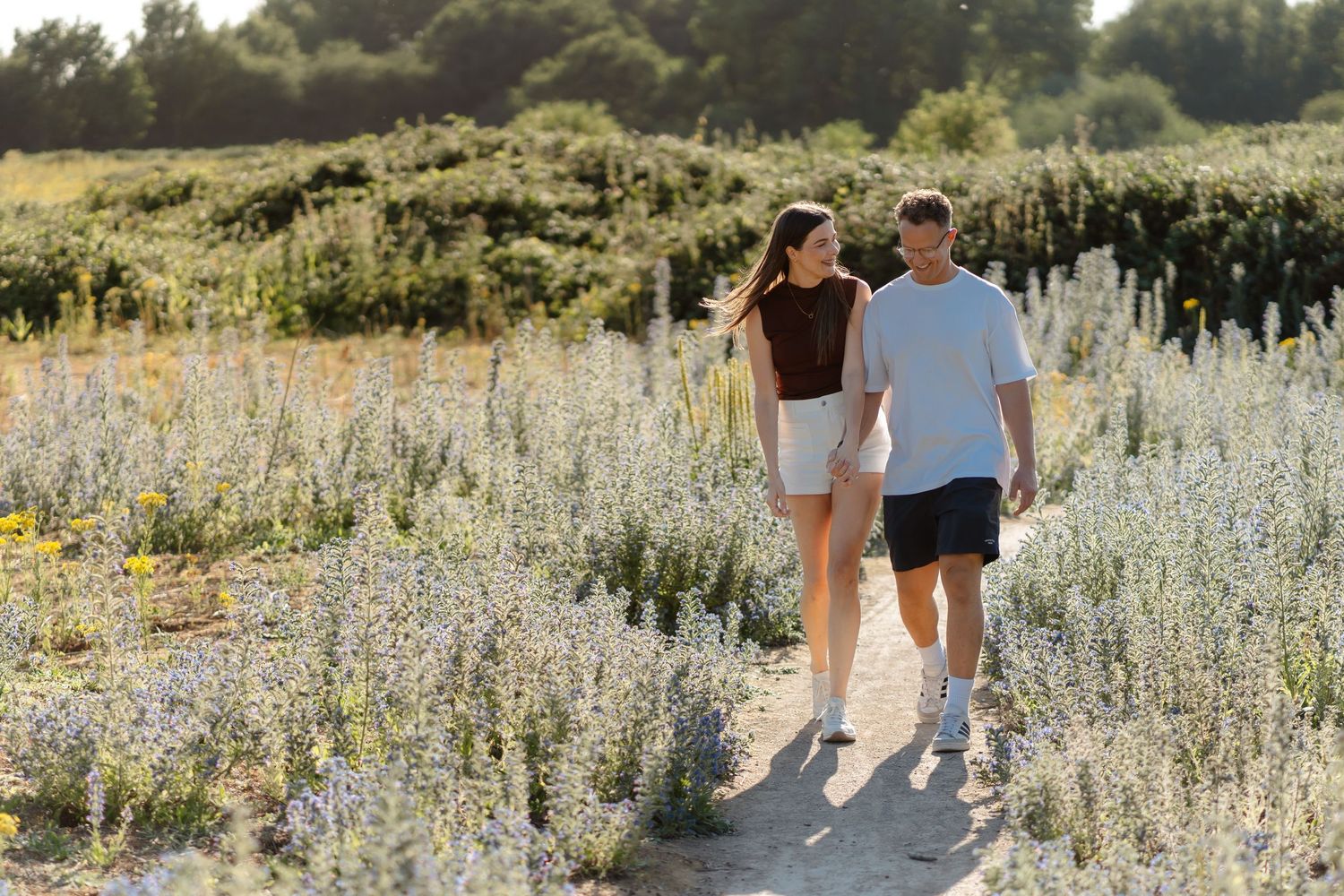 Couple holding hands walking on a sandy path through blooming wildflower fields on a sunny day.
