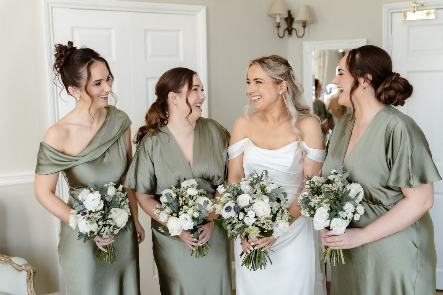 Bride and four bridesmaids in sage green dresses laughing together holding white floral bouquets before the wedding ceremony.