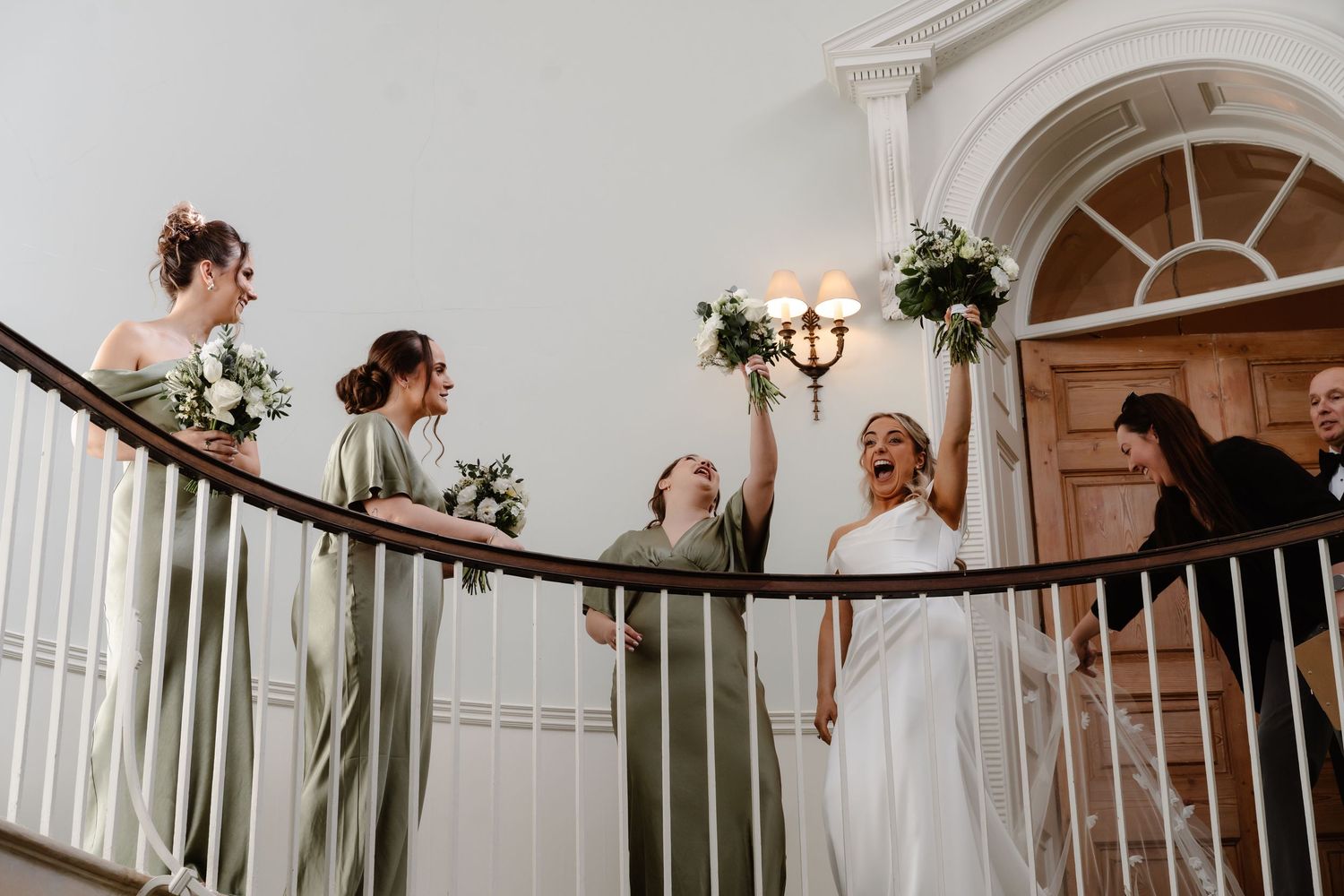 Bride and bridesmaids in sage green dresses celebrating on grand staircase with bouquets.