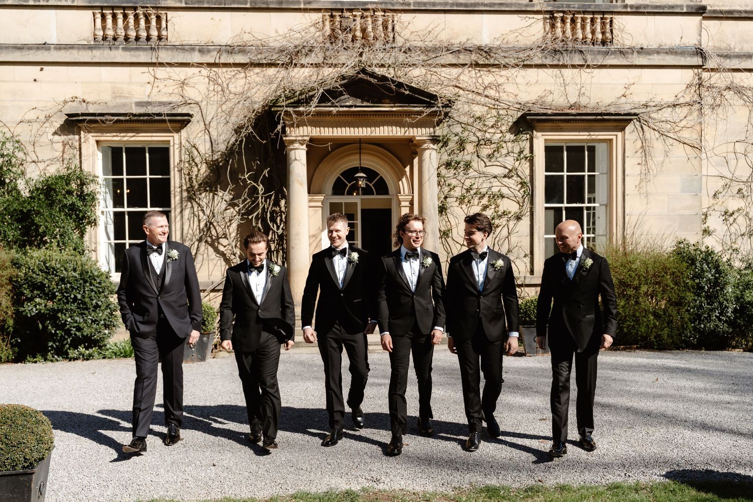 Groom and five groomsmen in black tuxedos walking confidently outside a grand stone estate.