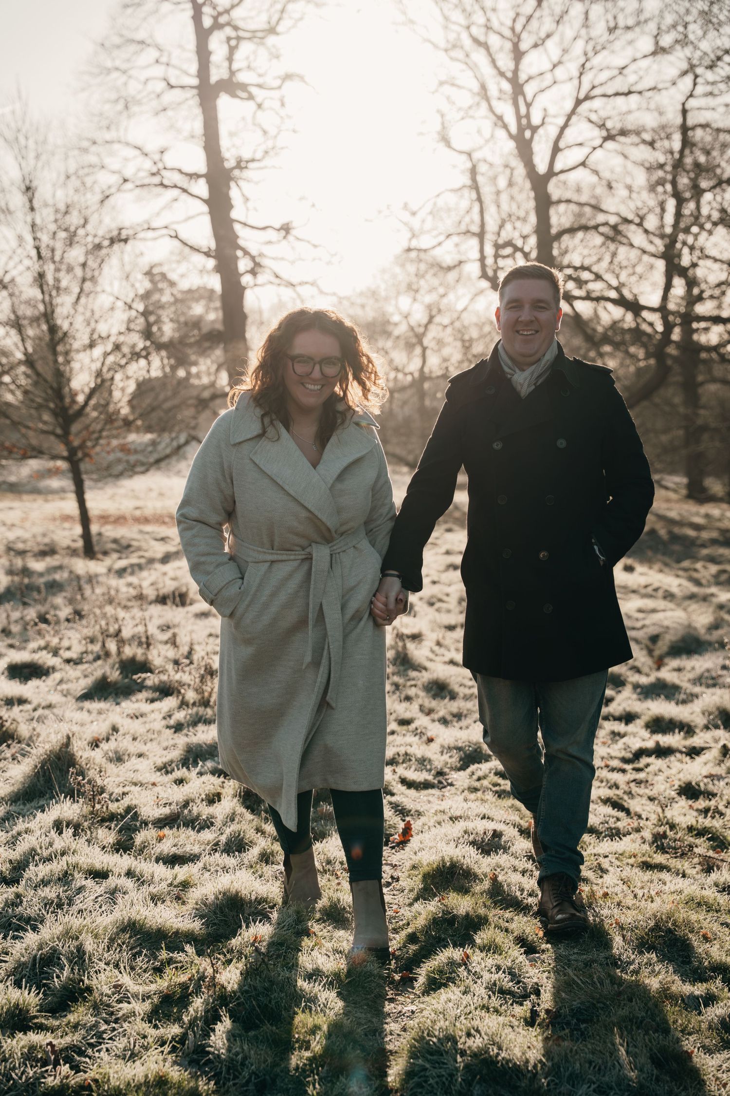 Couple holding hands walking through a frost-covered park in winter coats at sunrise.