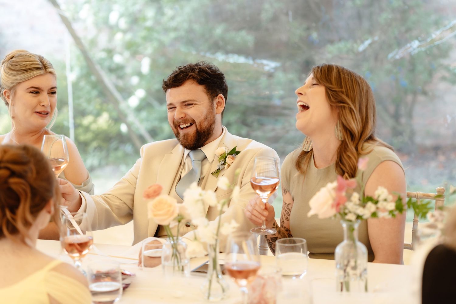 Bride and groom laughing with bridesmaid at elegant wedding reception table with floral centerpieces and wine glasses.