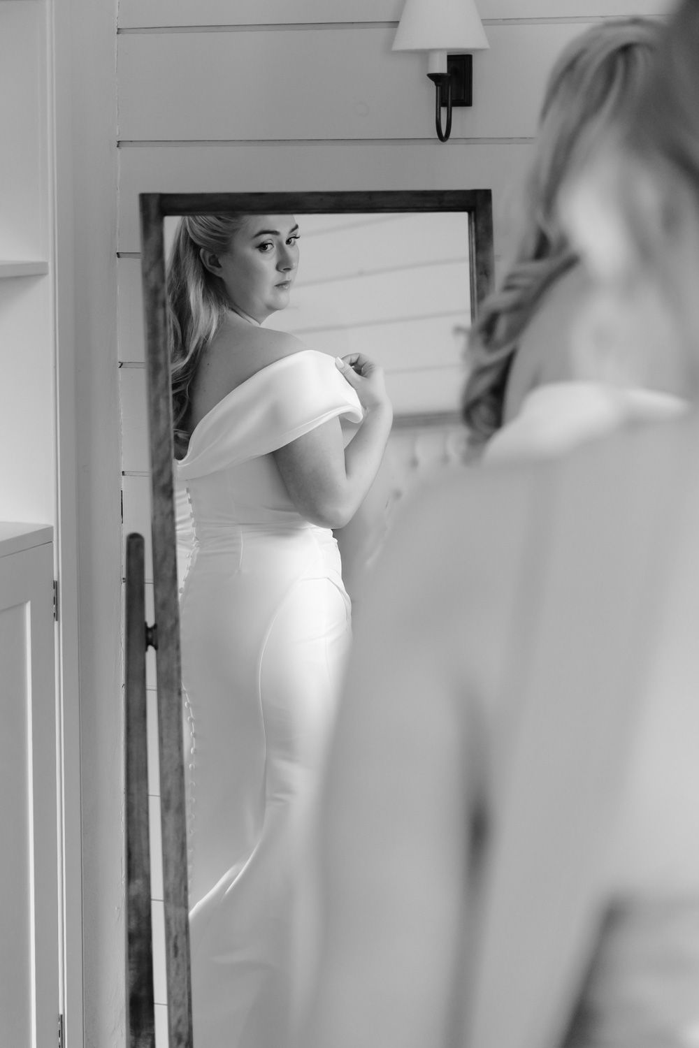 Bride in elegant off-shoulder gown checking her look in a full-length mirror.