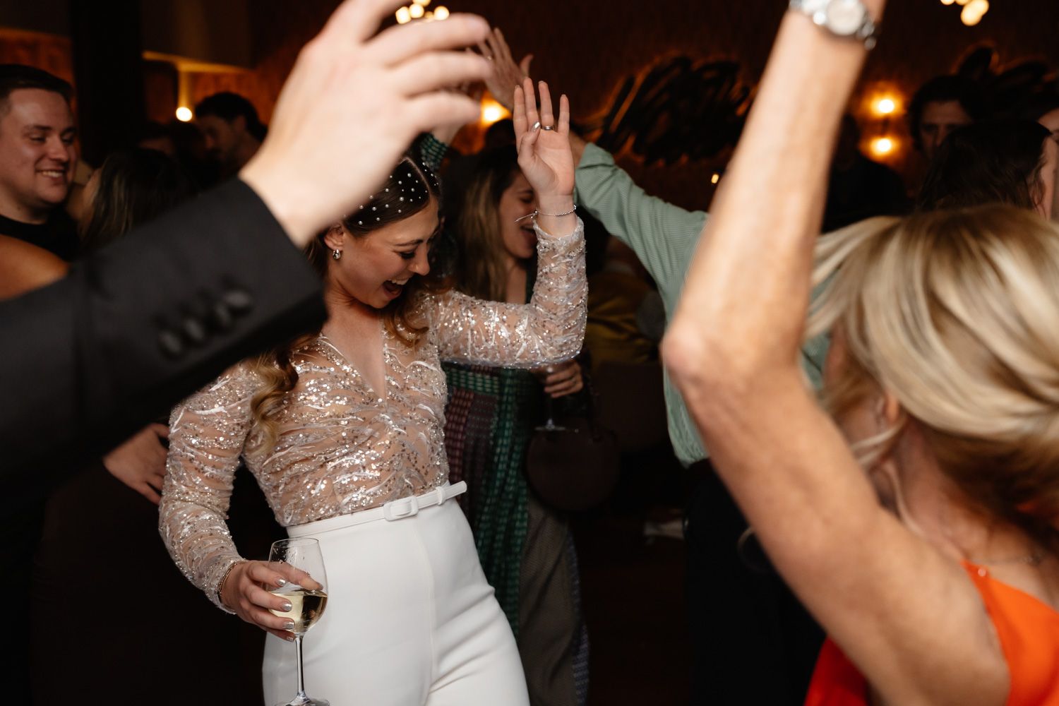 Bride in sequined top and white pants dancing joyfully at wedding reception.