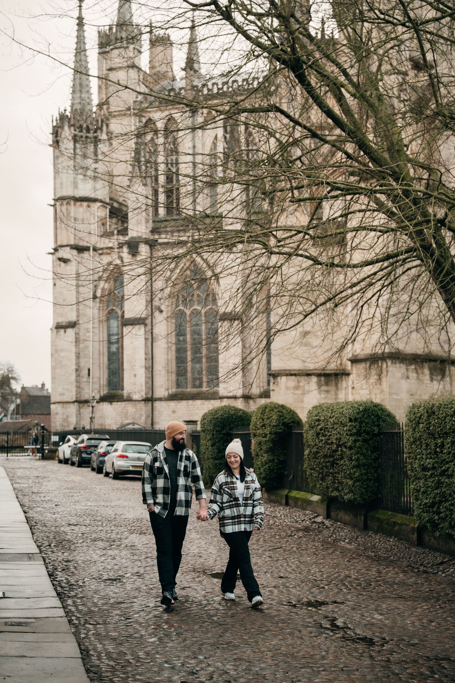 A couple in matching plaid shirts walk hand in hand along a cobblestone path beside a Gothic cathedral.