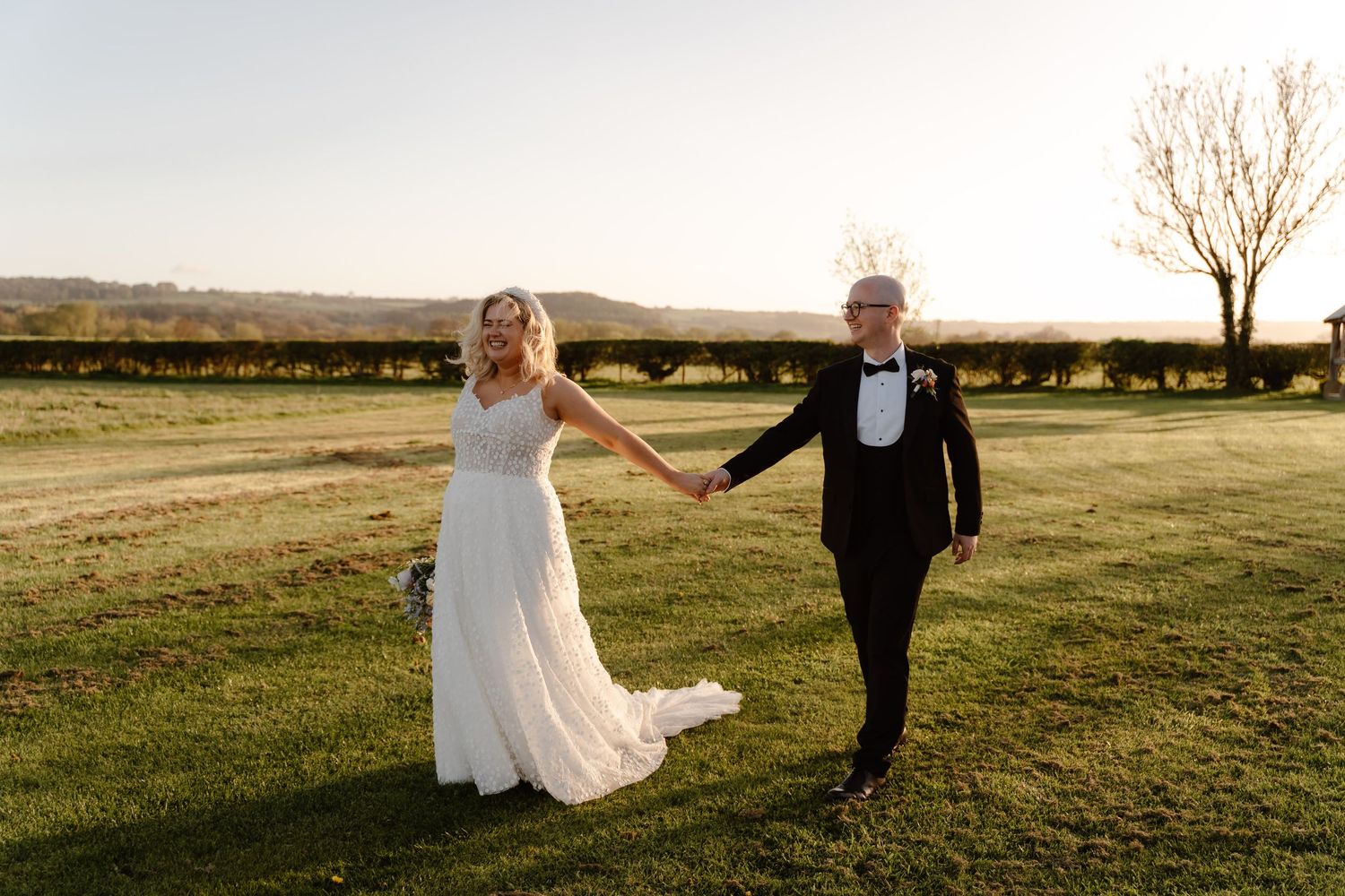 Bride and groom holding hands walking on green grass at golden hour sunset during outdoor wedding celebration.