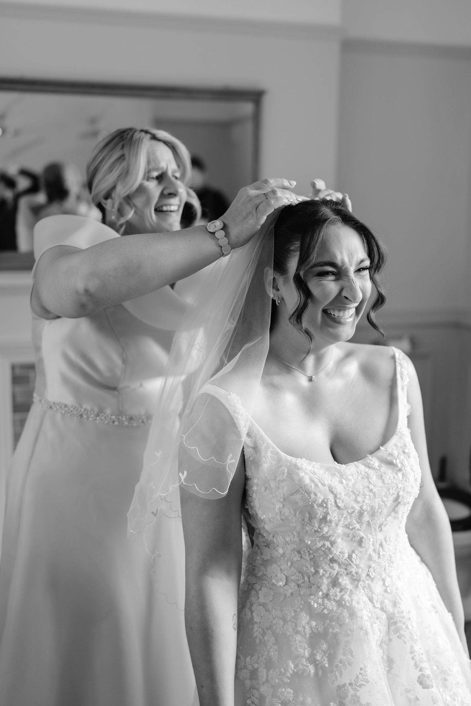Black and white photo of a bride smiling as her mother places a veil on her head during wedding preparations.