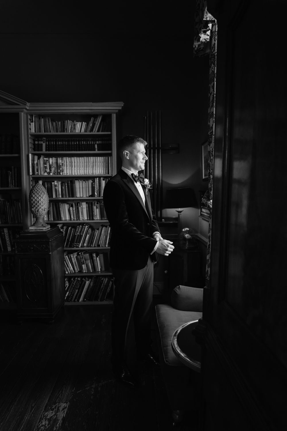 Black and white portrait of a groom in a tuxedo standing in an elegant library room.