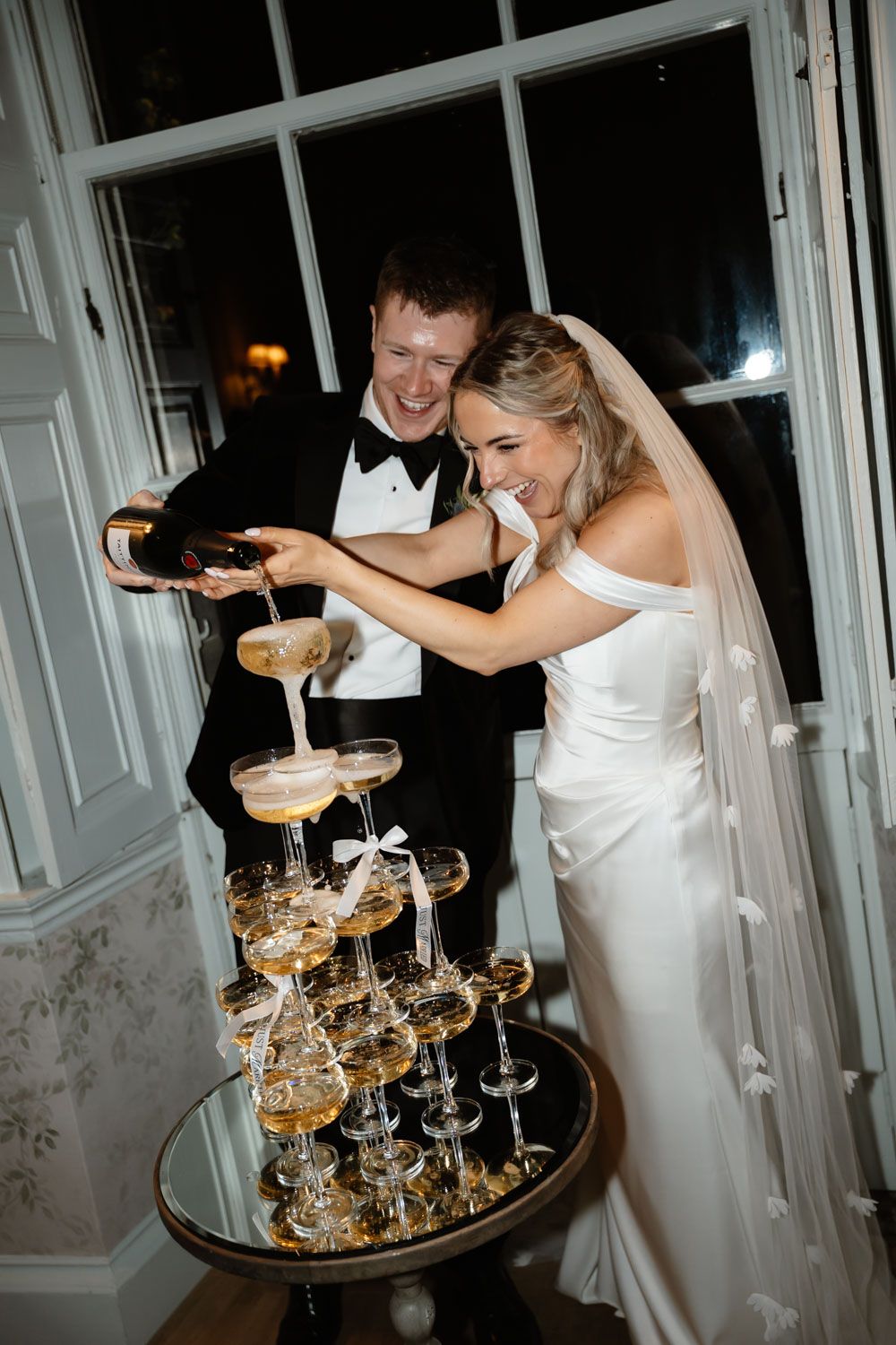 Smiling bride and groom pouring champagne over an elegant tiered coupe glass tower at reception.