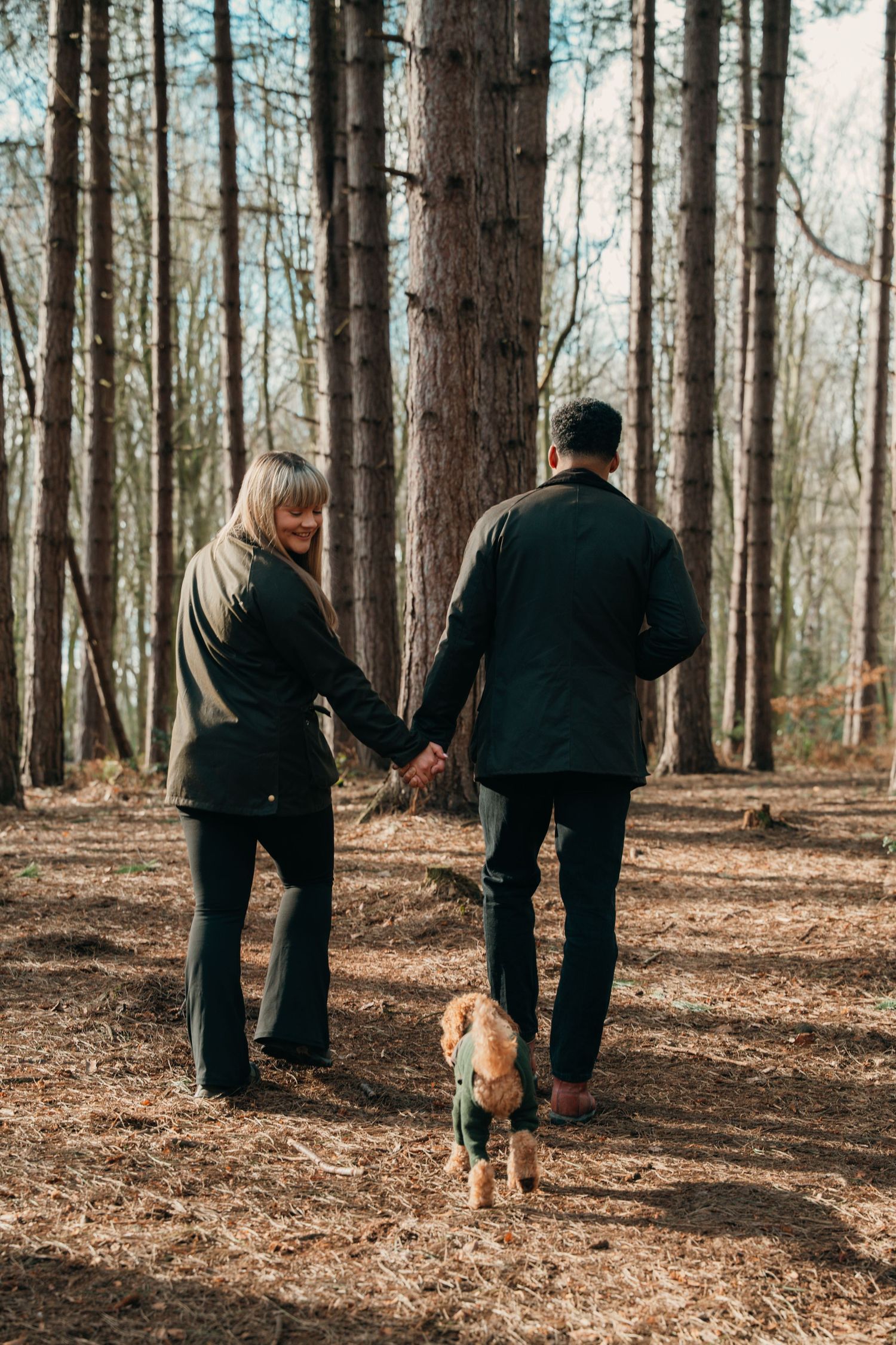 Couple holding hands walking away through woodland with small dog in green jacket.