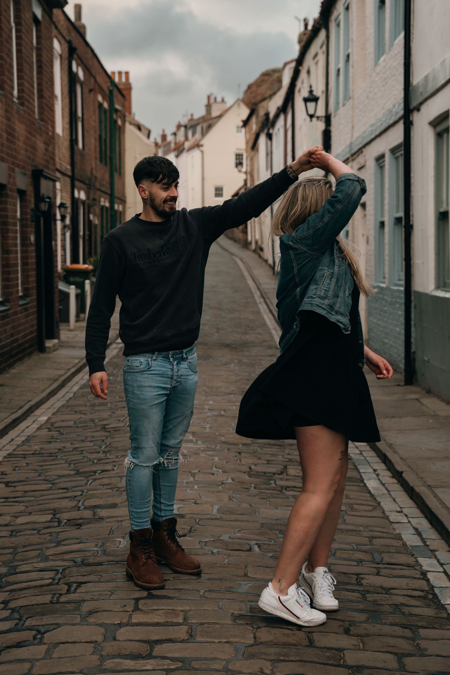 Couple dancing on cobblestone street in historic town, man spinning woman in black skirt and denim jacket.