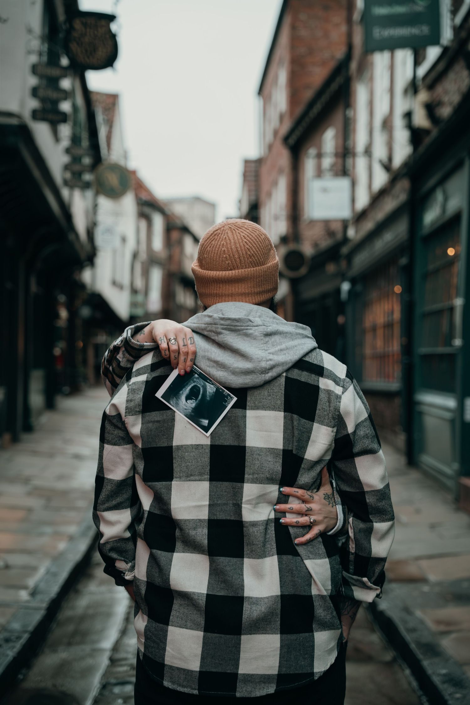 A man in a plaid shirt and beanie holds a baby ultrasound scan on a historic cobblestone street.